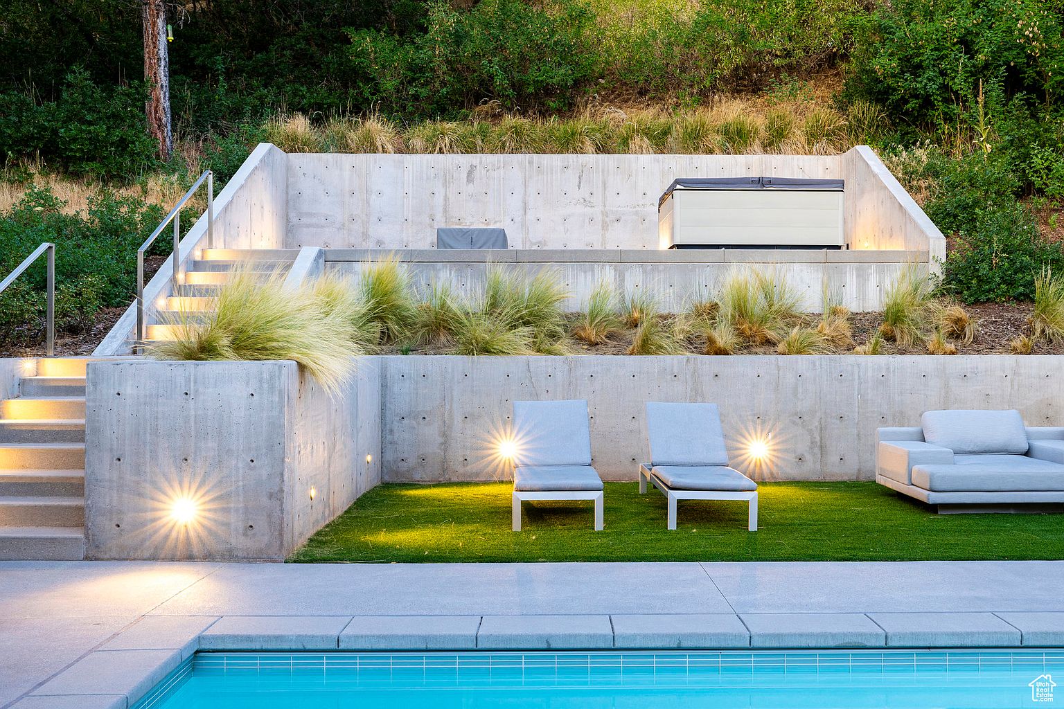 This image showcases a modern backyard oasis featuring a sleek pool with a concrete surround. The tiered landscaping includes concrete retaining walls, ornamental grasses, and a hot tub on the upper level. Lounge chairs and a sofa provide ample seating for relaxation, creating an inviting and stylish outdoor living space.