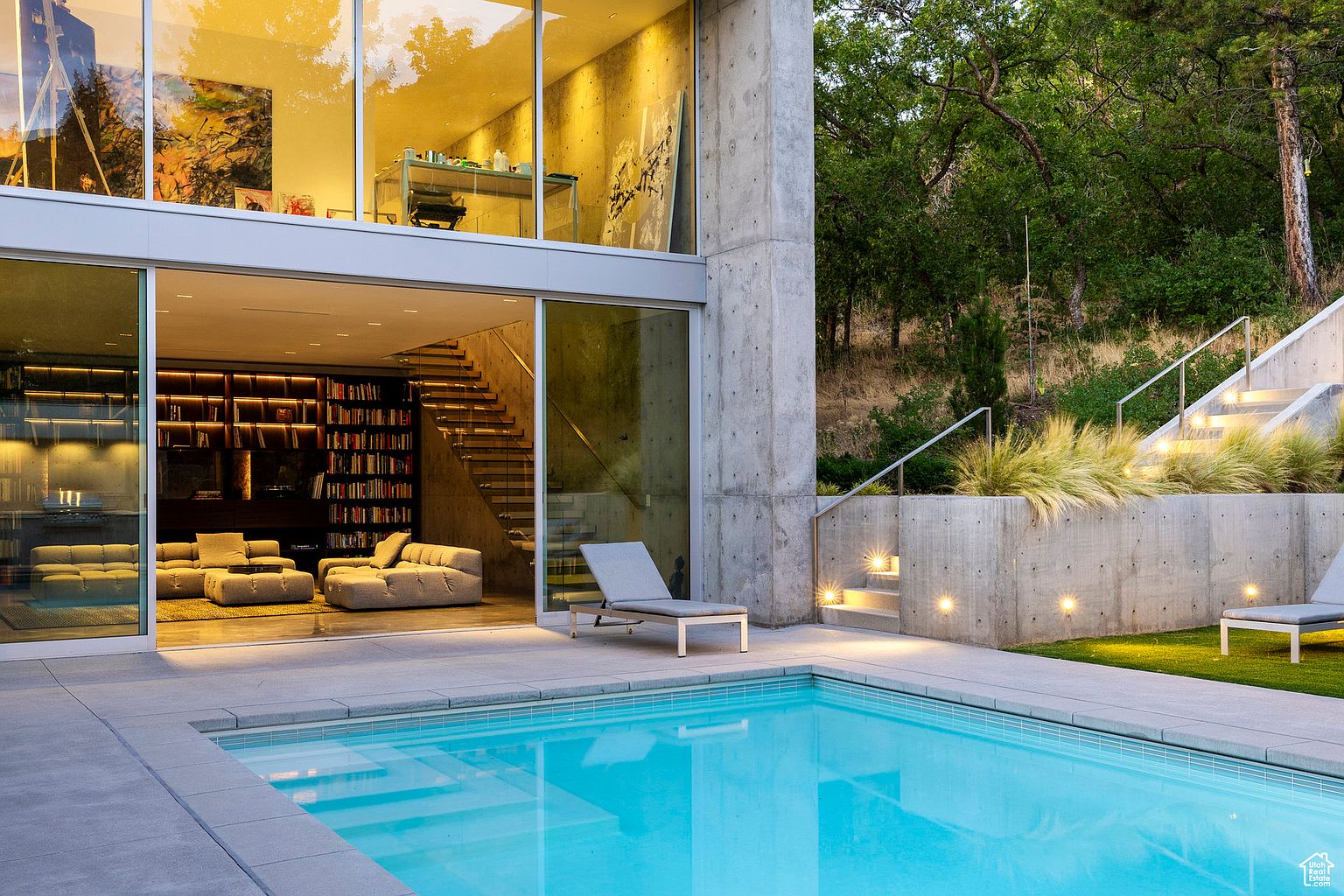 This image showcases a modern home's exterior with a focus on the pool area. The pool features clear blue water and is surrounded by a concrete patio. Large glass doors open to the home's interior, revealing a living space with a bookshelf and stairs, while a concrete staircase leads up the hillside, complemented by landscape lighting and greenery.