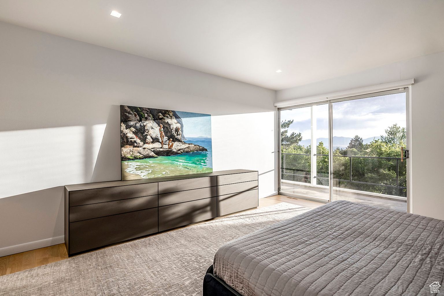 This is a bright and airy primary bedroom featuring a large sliding glass door that leads to a balcony with a view of the surrounding trees and mountains. The room is furnished with a low dresser with a large painting above it and a bed with a gray quilted cover. The flooring is light wood, and a neutral-toned rug covers the floor.