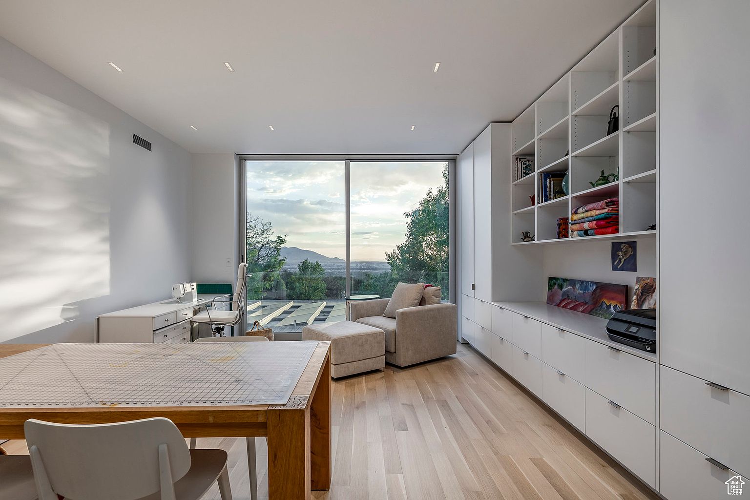 This is a bright and modern office space featuring a large window with a scenic view. The room includes built-in shelving and cabinets, a comfortable armchair with an ottoman, and a spacious work table. The light wood flooring and minimalist design create a clean and inviting atmosphere, perfect for a home office or study.