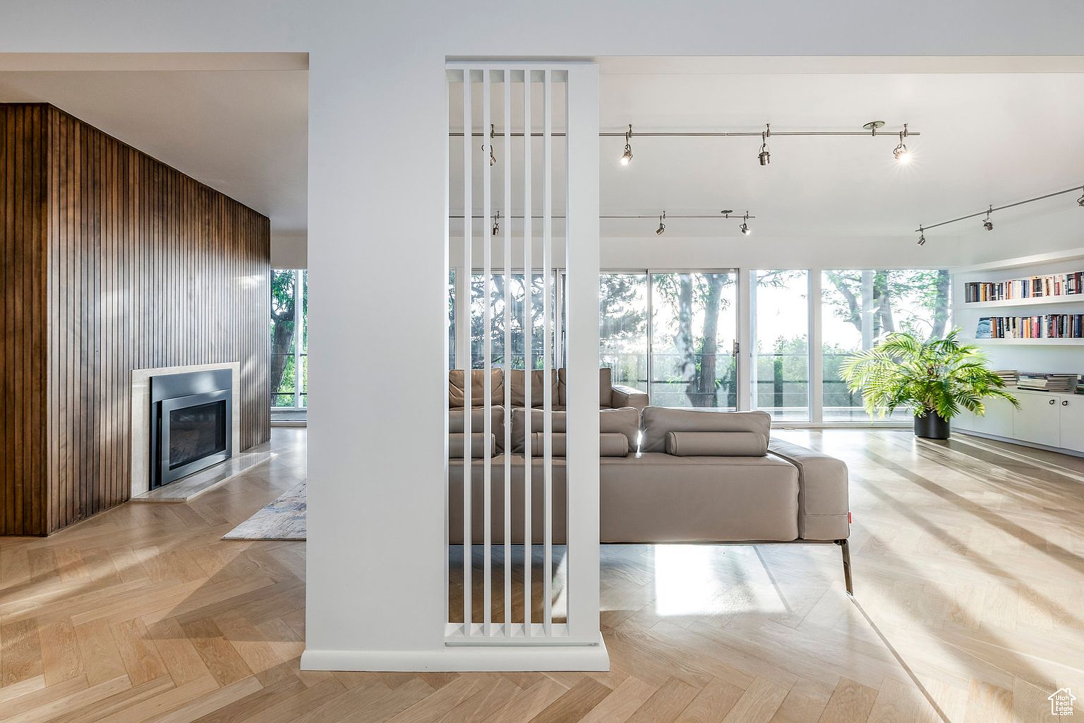 This is a bright and spacious living room featuring a modern design with clean lines and a neutral color palette. The room is divided by a stylish white partition, separating the seating area from a wood-paneled wall with a fireplace. Large windows provide ample natural light and views of the surrounding greenery, enhancing the open and airy feel of the space.