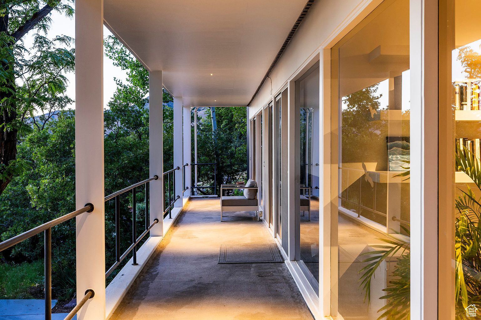 This image showcases a covered balcony or patio area, featuring white support columns and a metal railing. The space is furnished with a modern outdoor seating arrangement, and large glass doors provide access to the interior. The view from the balcony includes lush greenery, creating a serene and inviting atmosphere.