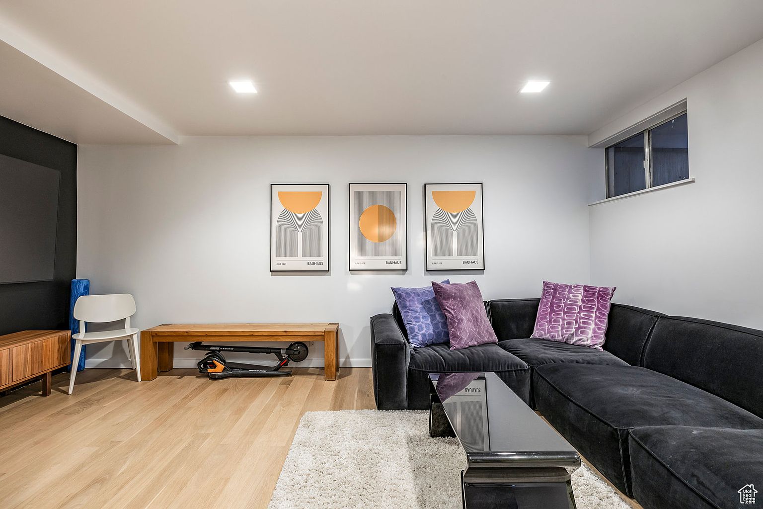 This is an interior shot of a modern living room featuring a dark gray sectional sofa with purple pillows, a black coffee table, and a light-colored shag rug. Three framed Bauhaus-style art prints hang on the wall above the sofa. The room has light wood flooring and recessed lighting, creating a bright and inviting space.