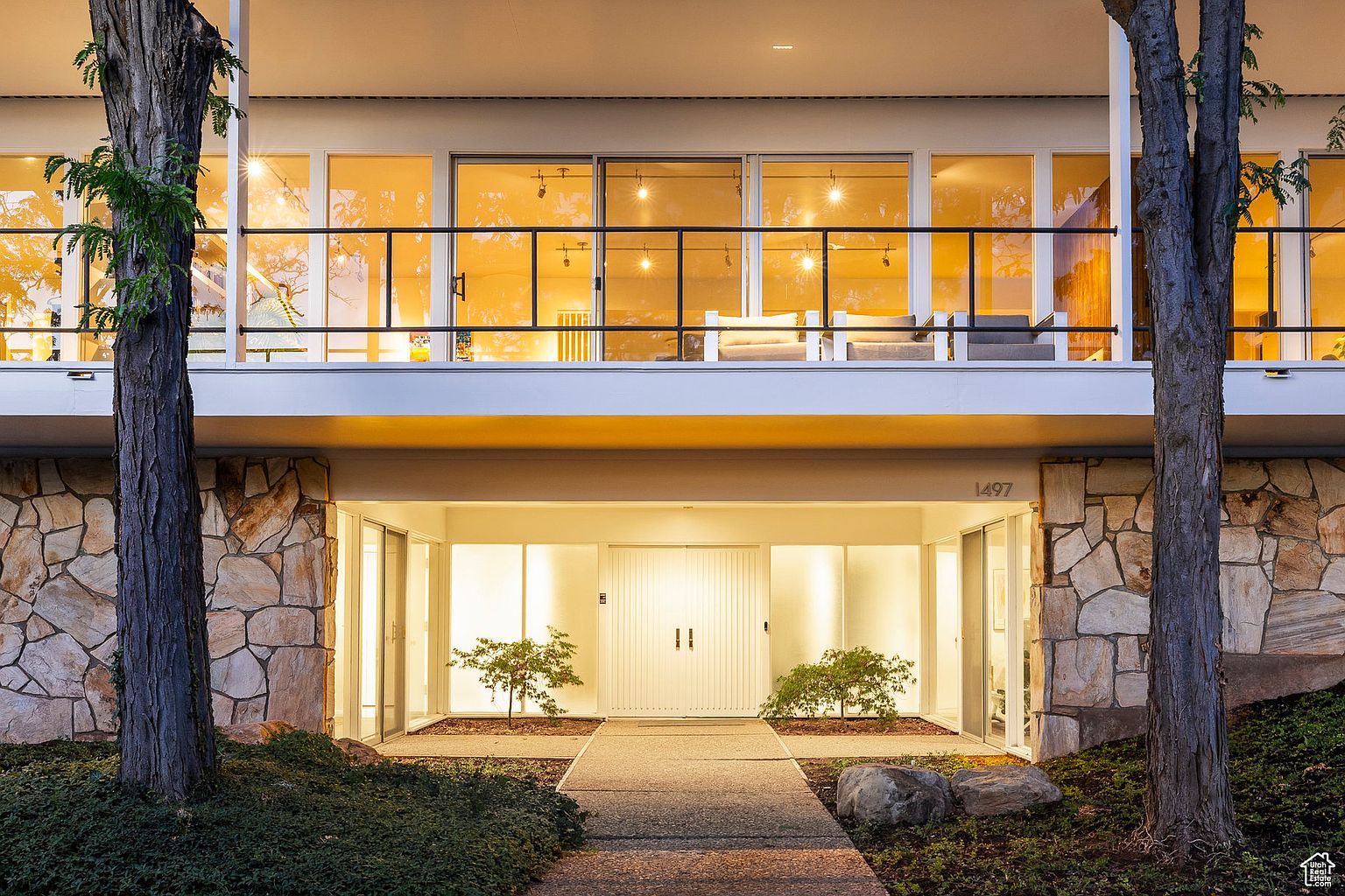 This image showcases the entryway of a modern home, featuring a stone facade on either side of a covered porch. The entrance includes a white double door, flanked by large glass panels that allow ample light into the interior. A paved walkway leads to the entrance, framed by trees and landscaping, creating an inviting and sophisticated first impression.
