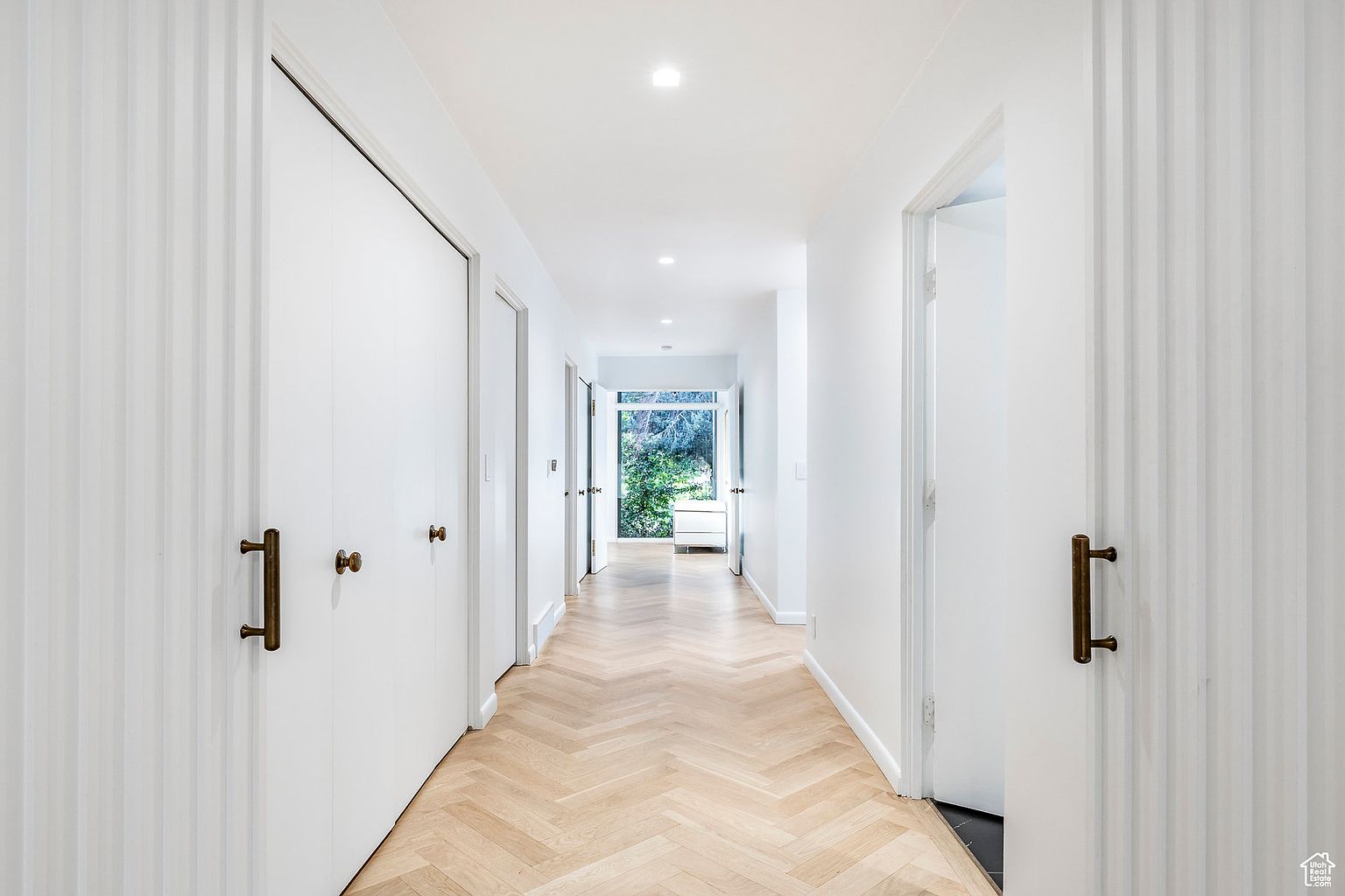 This is a bright and modern hallway featuring white walls, doors, and trim, complemented by a light wood herringbone floor. The hallway is well-lit with recessed lighting and natural light streaming in from the end, creating a clean and inviting atmosphere. The perspective is a straight-on view down the hallway, emphasizing its length and the transition to the outdoor view at the end.