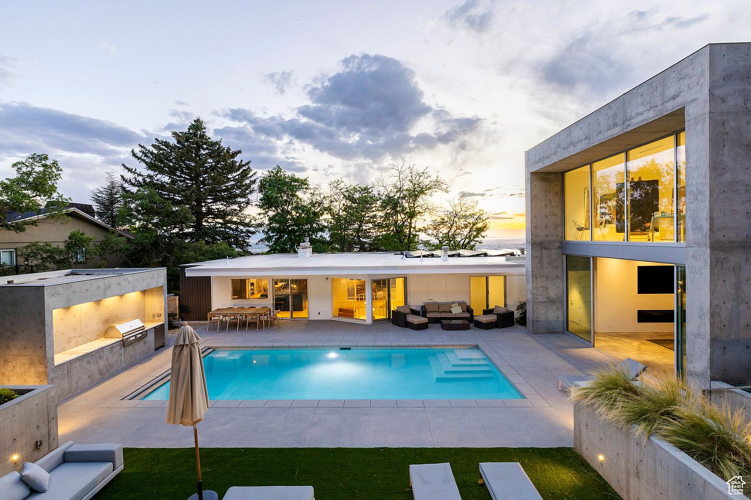 This image showcases a luxurious backyard with a modern aesthetic. A rectangular pool takes center stage, surrounded by a concrete patio and a patch of artificial grass. The property features a contemporary home with large windows and a concrete structure, complemented by outdoor seating and a built-in grill area, creating an inviting space for relaxation and entertainment.