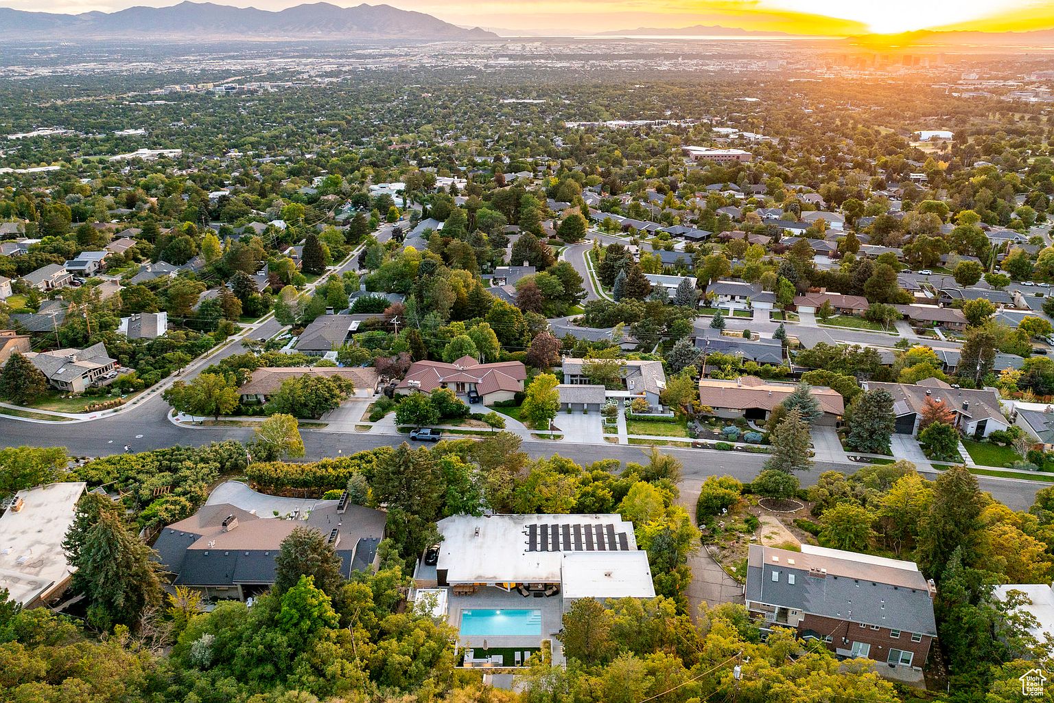 This aerial view showcases a residential neighborhood with lush greenery and well-maintained homes. A standout property features a modern design with a pool and solar panels, nestled among mature trees. The landscape extends to a distant mountain range under a warm, sunset-lit sky, creating a serene and desirable setting.