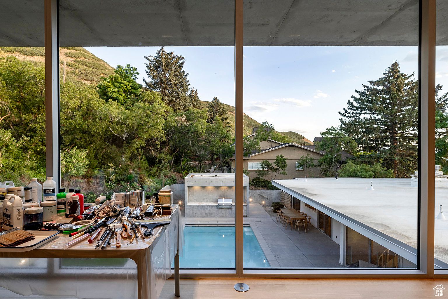 This image showcases a modern home's exterior, specifically the pool and patio area, as seen through a large window. The pool is surrounded by a concrete patio with outdoor dining furniture, and a built-in grill area is visible. Lush greenery and trees provide a scenic backdrop, enhancing the property's appeal and creating a sense of privacy and tranquility.