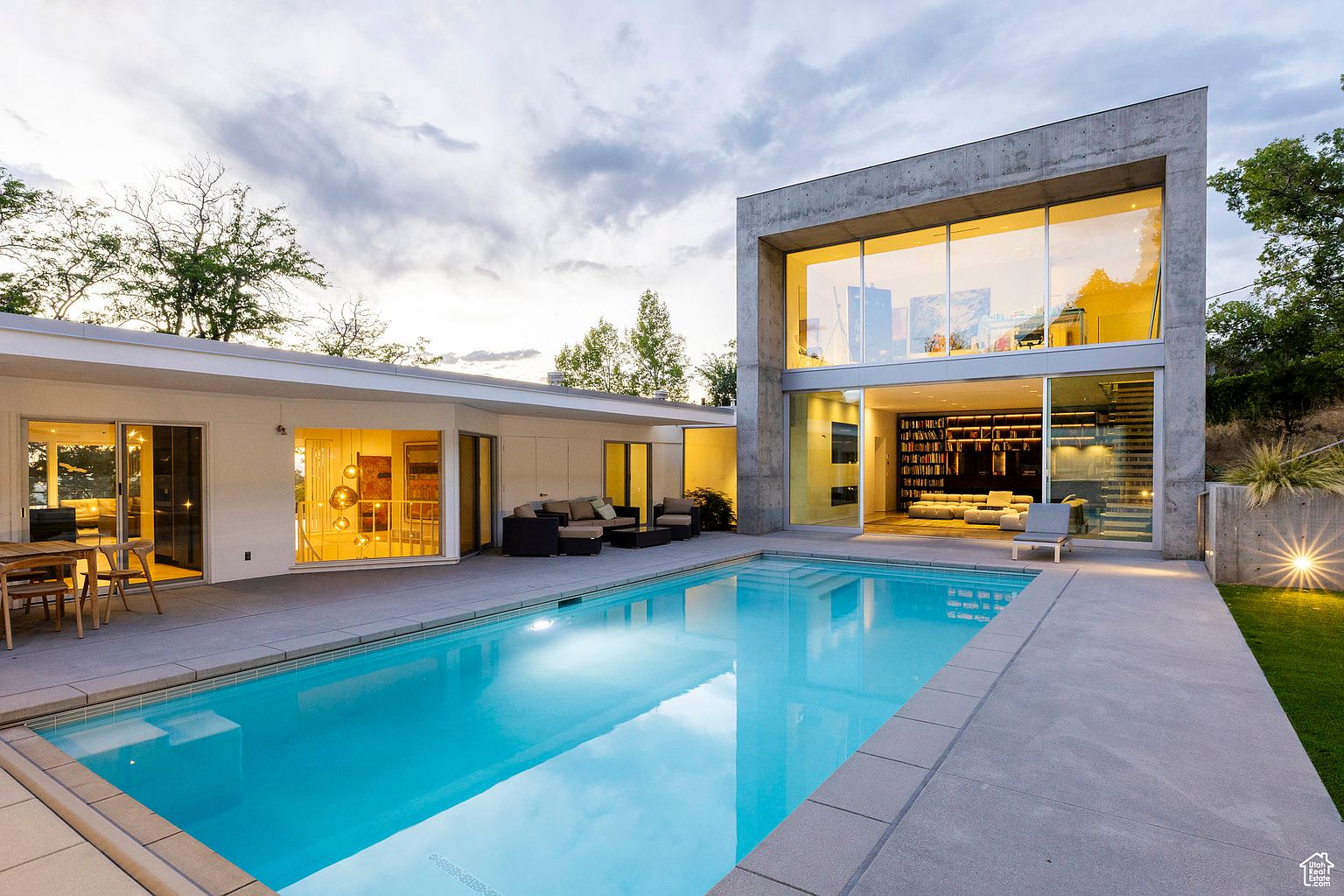 This image showcases a modern home's exterior with a focus on the pool area. The pool's clear blue water reflects the sky, flanked by a concrete patio. The architecture features a striking concrete structure with large windows and a connecting white building, creating a luxurious and inviting outdoor space.