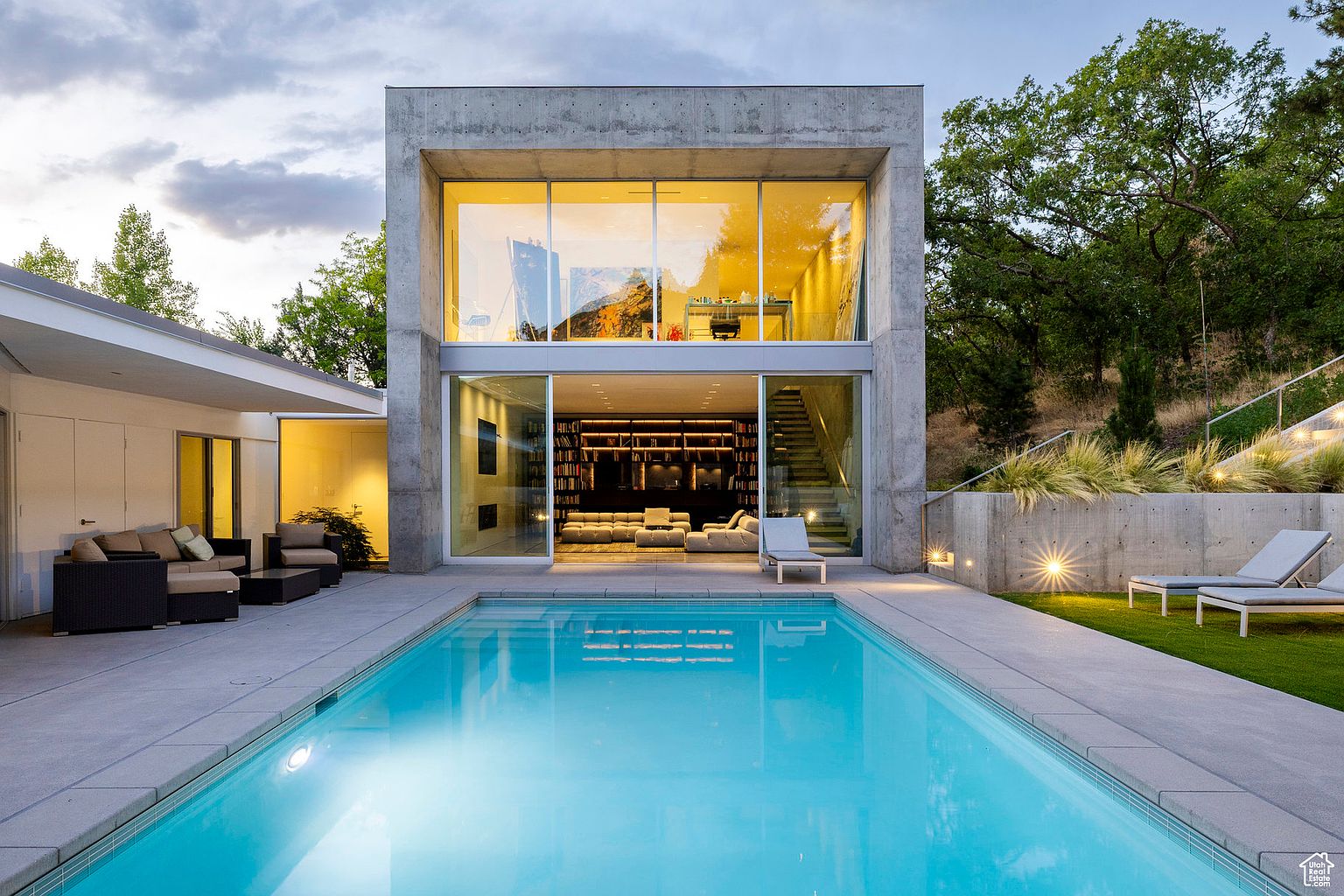 This image showcases a modern home's exterior with a focus on the pool area. The rectangular pool is surrounded by a concrete patio, leading to a two-story concrete structure with large glass windows and doors. The architecture is minimalist and contemporary, with lush greenery in the background, creating a luxurious and serene atmosphere.