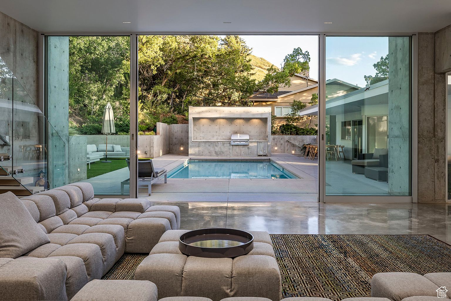 This image showcases a modern living room that seamlessly blends indoor and outdoor spaces. The room features a large, modular sofa in a neutral tone, complemented by a geometric rug. Floor-to-ceiling glass doors open to a backyard with a pool and outdoor kitchen, creating an inviting and luxurious atmosphere.
