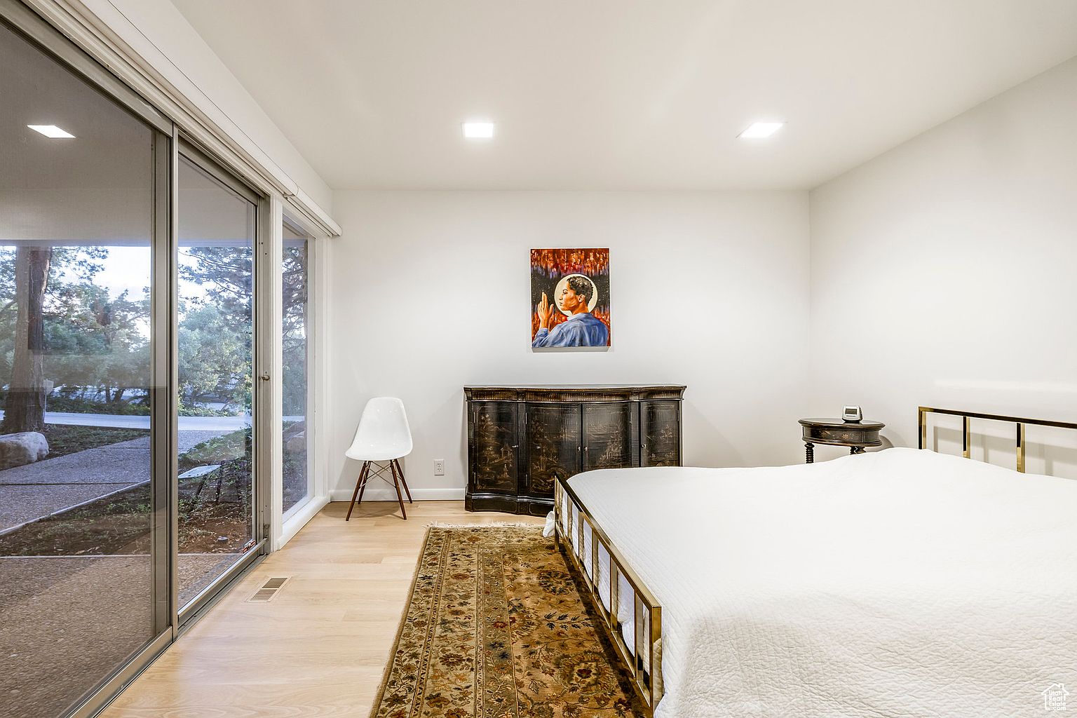 This is a bright and airy primary bedroom featuring a large sliding glass door that provides ample natural light and a view of the outdoors. The room is minimally decorated with a white bed, a dark wood dresser, and a piece of artwork above it. A neutral-toned rug adds warmth to the light wood flooring, creating a serene and inviting atmosphere.