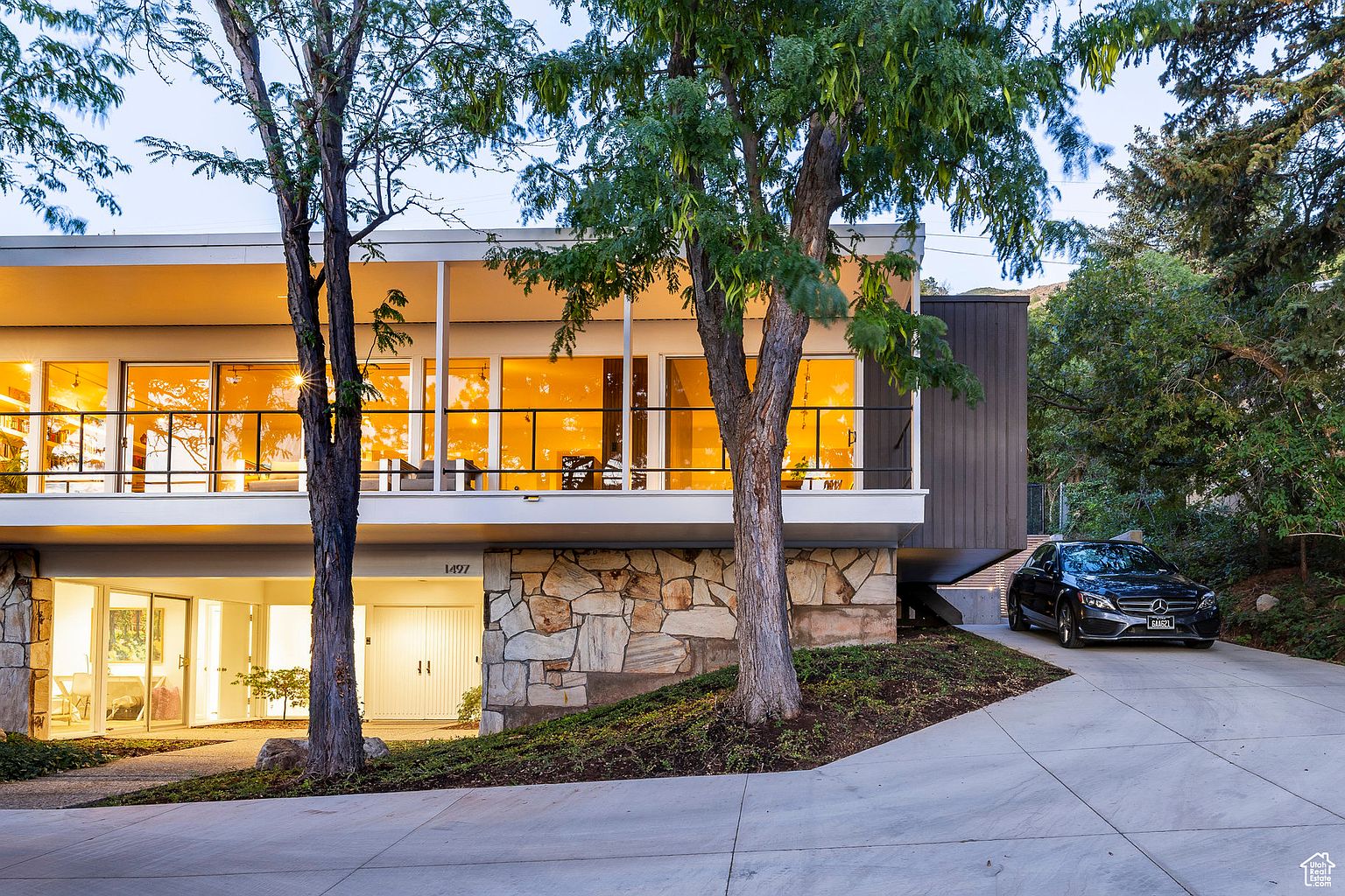 This is a front exterior view of a modern, two-story house. The upper level features a wall of windows with a balcony, while the lower level is constructed with stone. A black car is parked in the driveway, and mature trees frame the house, adding to its curb appeal.