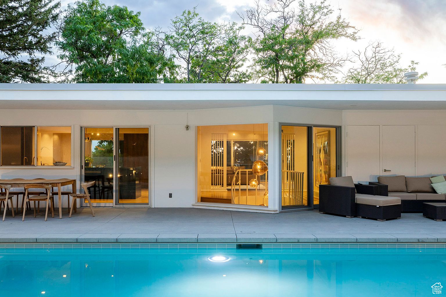 This image showcases a modern home's exterior with a focus on the pool area. The pool's clear blue water reflects the sky, and a patio with outdoor furniture extends from the house. Large windows and sliding glass doors provide seamless indoor-outdoor living, enhancing the property's appeal.