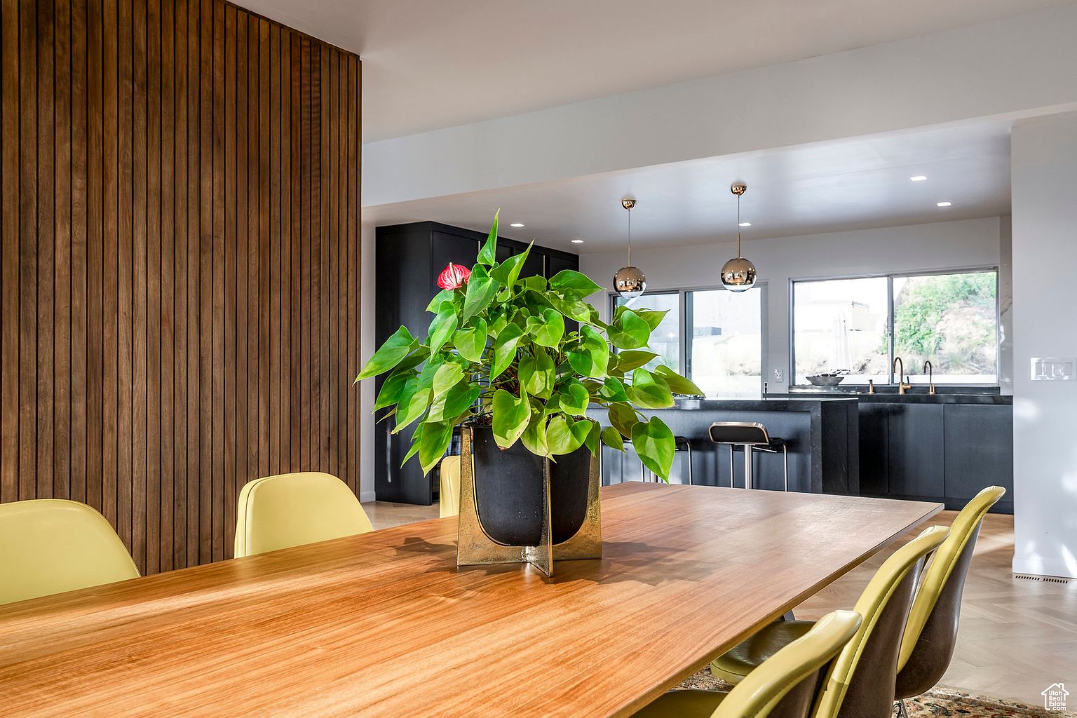 This interior shot showcases a dining room with a wooden table as the focal point, adorned with a vibrant green plant in a decorative pot. The room features modern design elements, including a wooden slat wall and stylish chairs, with a glimpse into a contemporary kitchen in the background. The overall impression is one of sophisticated comfort and modern living.