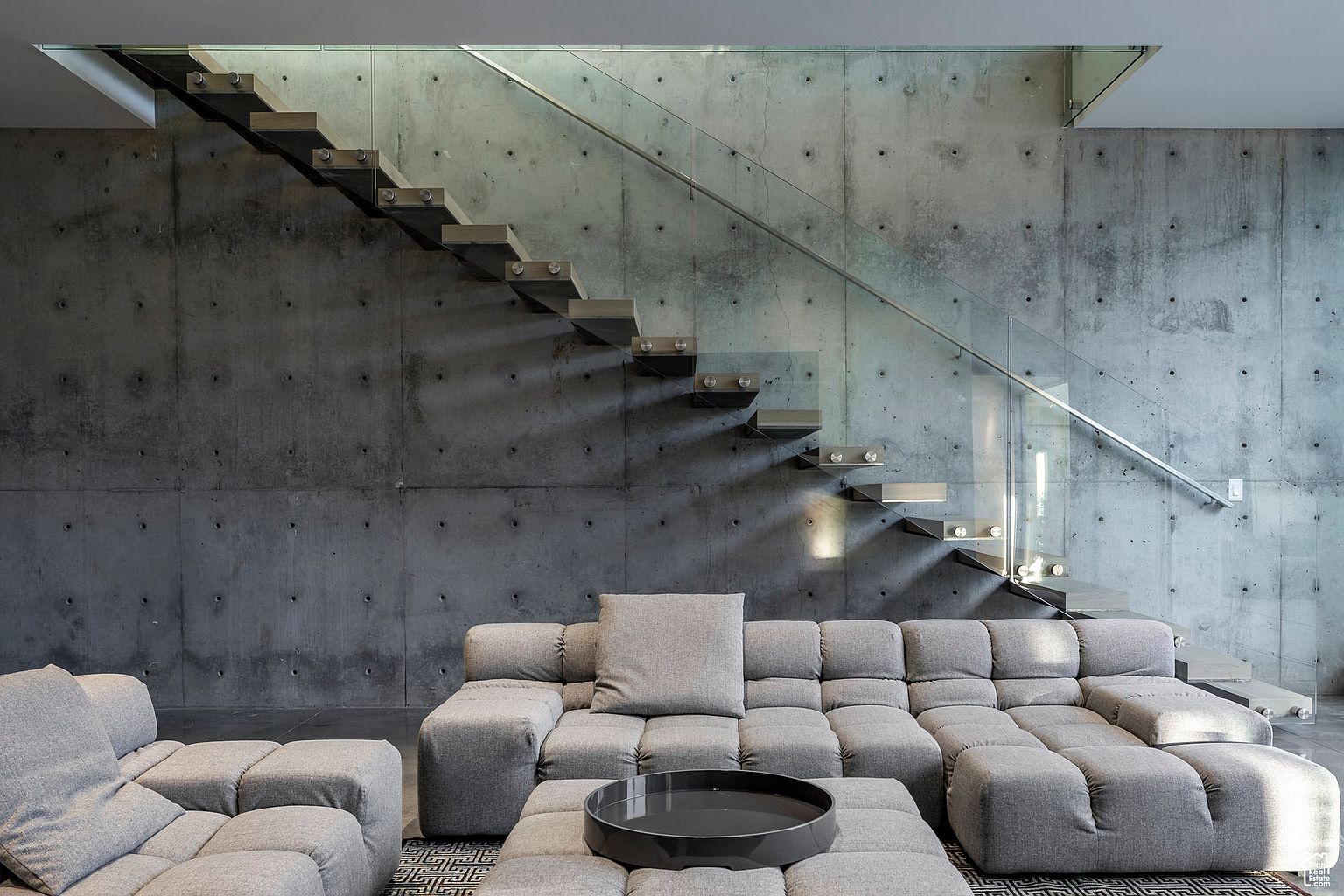This interior shot showcases a modern living room with a striking concrete wall and floating staircase with glass railings. A large, modular gray sofa provides ample seating, complemented by a matching ottoman and a sleek, dark-colored tray. The room exudes a minimalist yet luxurious aesthetic, emphasizing clean lines and textural contrast.