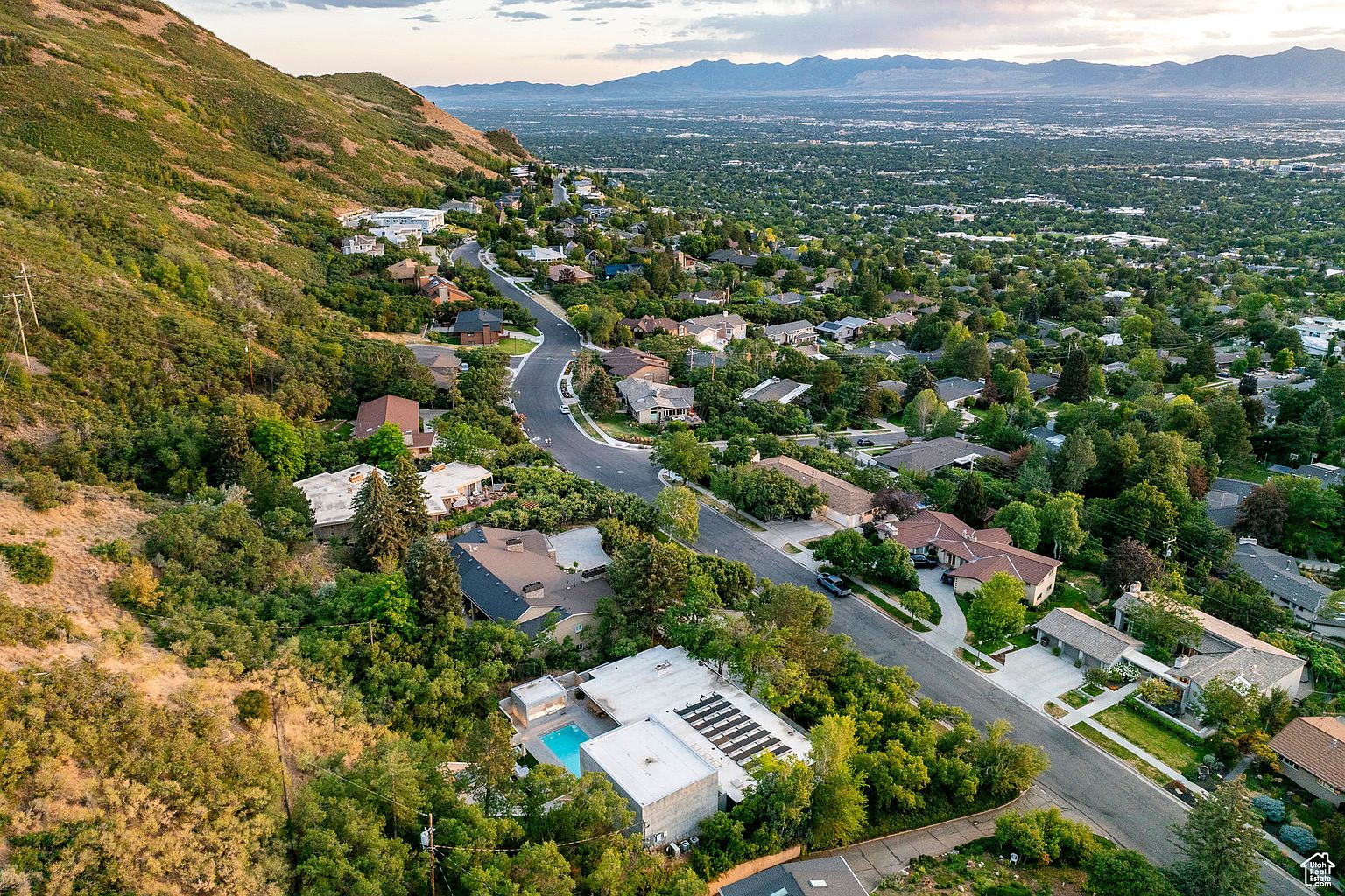 This aerial view showcases a neighborhood nestled on a hillside, blending residential architecture with lush greenery. The winding roads connect various homes, some featuring pools and solar panels, against a backdrop of distant mountains and a sprawling cityscape. The perspective gives a sense of community and natural integration.