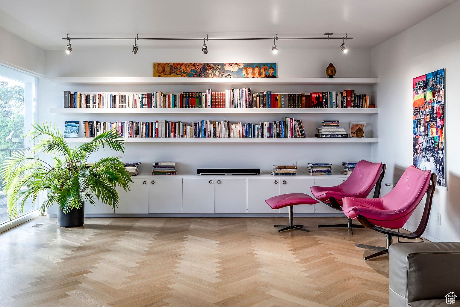 This is an interior shot of a living room featuring a herringbone wood floor, a large built-in bookshelf filled with books and decorative items, and two pink lounge chairs with a matching ottoman. The room is well-lit with track lighting and natural light from a nearby window, creating a bright and inviting atmosphere. A modern art piece hangs on the wall to the right.