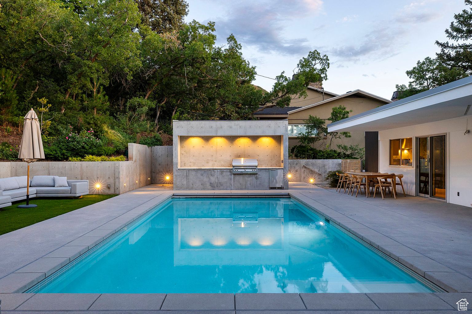 This image showcases a modern backyard with a rectangular swimming pool as the central feature. The pool is surrounded by a concrete patio, with a built-in outdoor kitchen area featuring a grill and lighting. A seating area with an umbrella and sofa is visible on a small patch of grass, and the yard is bordered by lush greenery, creating a private and luxurious outdoor living space.