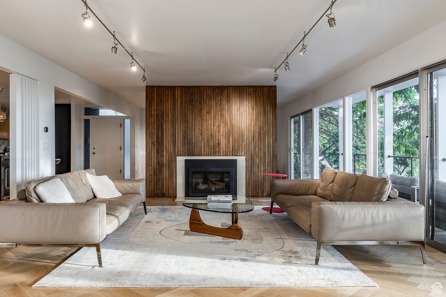 This is a well-lit living room featuring two modern leather sofas facing each other, separated by a glass-topped coffee table. A striking wood-paneled wall with a fireplace serves as a focal point, complemented by track lighting on the ceiling. Large windows provide natural light and a view to the outside, enhancing the room's spacious feel.