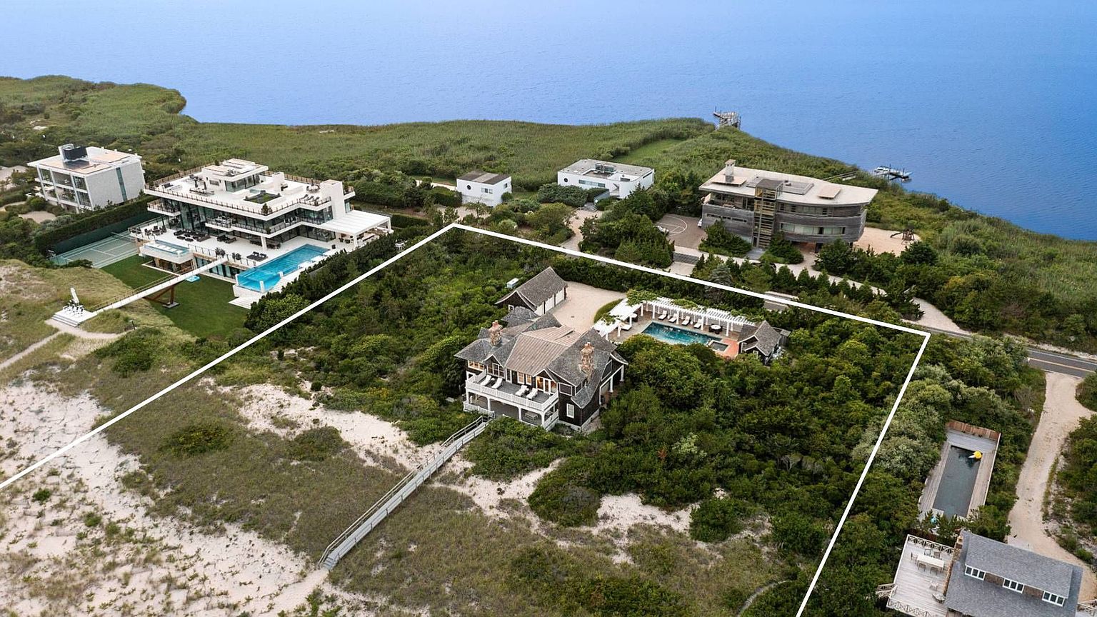 This aerial shot showcases a luxurious estate with a sprawling property, including a large house with a dark roof, a pool, and a walkway leading to the beach. The property is surrounded by lush greenery and is situated near the water, offering stunning views and a sense of privacy. The image highlights the scale and exclusivity of the real estate.