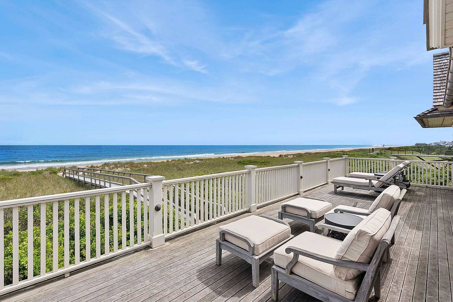 This image showcases a spacious wooden deck overlooking a beautiful beach and ocean. The deck is furnished with comfortable lounge chairs and ottomans, creating an inviting outdoor living space. A white railing surrounds the deck, providing safety and enhancing the coastal aesthetic.