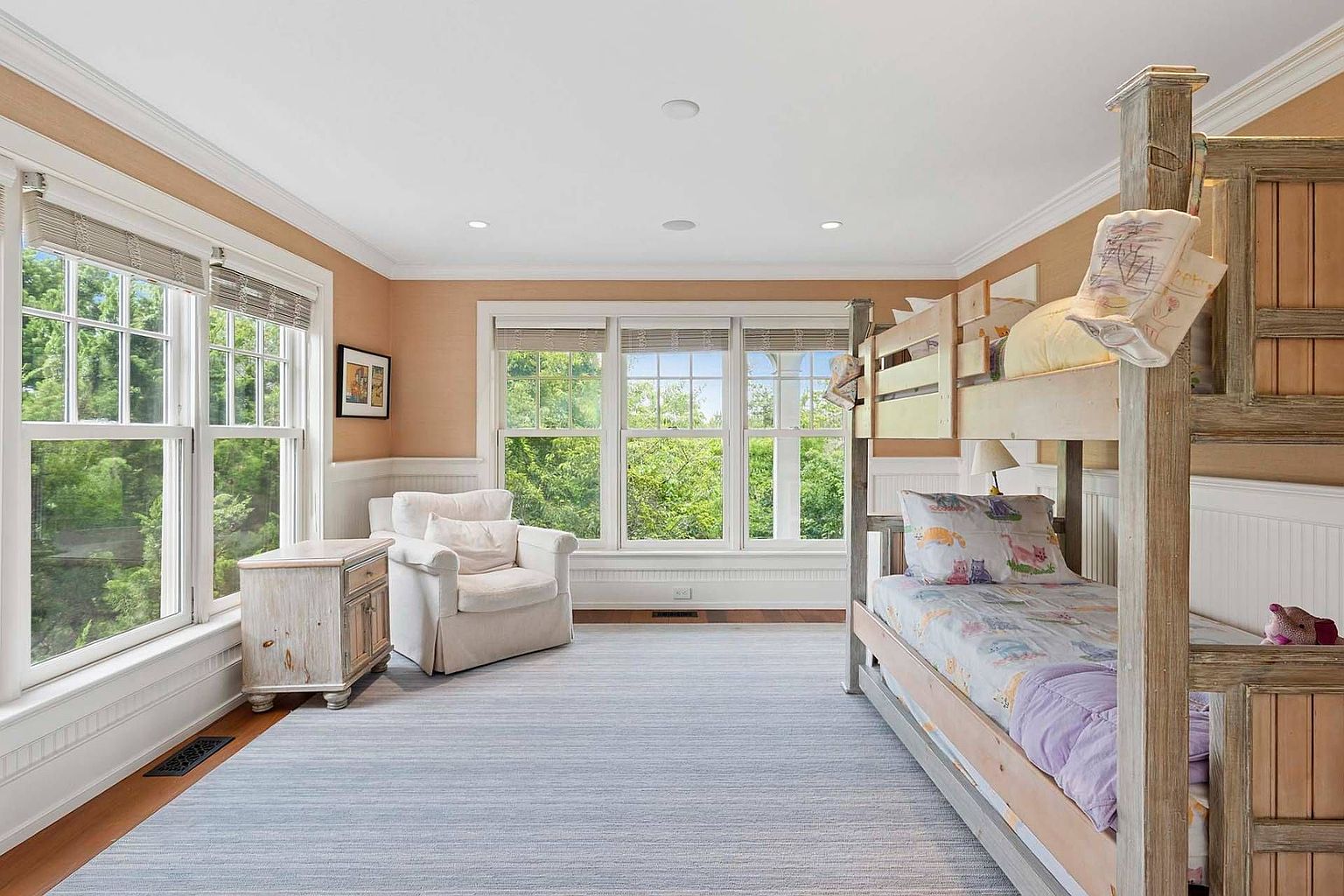 This is a bright and airy bedroom featuring a bunk bed, a comfortable armchair, and a large window offering a view of lush greenery. The room is decorated in a neutral color palette with light blue accents, creating a calming and inviting atmosphere. The hardwood floors and white wainscoting add a touch of elegance to the space.