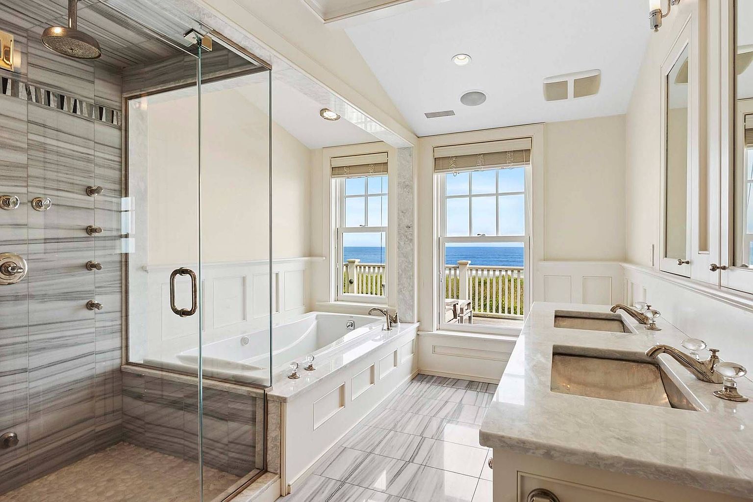 This is a luxurious primary bathroom featuring a glass-enclosed shower with multiple showerheads and marble tiling. A soaking tub is situated beneath two windows offering an ocean view. The bathroom also includes a double vanity with marble countertops and elegant fixtures, creating a spa-like atmosphere.