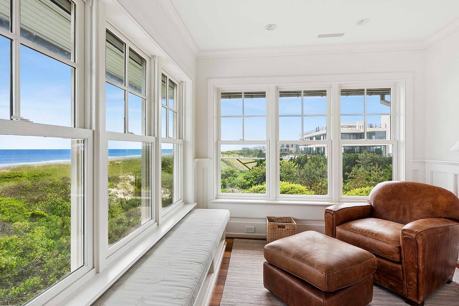This sunroom features large windows offering expansive views of the beach and surrounding landscape. A comfortable leather chair and ottoman create a cozy seating area, complemented by a woven basket and a cushioned window seat. The room is bright and airy, with white trim and walls enhancing the natural light.