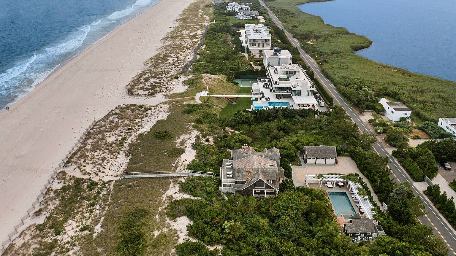 This aerial shot showcases a stunning coastal property featuring a mix of modern and traditional architectural styles. The homes are nestled amidst lush greenery, with direct access to a pristine sandy beach on one side and a tranquil body of water on the other. The presence of pools, tennis courts, and well-maintained landscaping adds to the luxurious appeal of these residences.