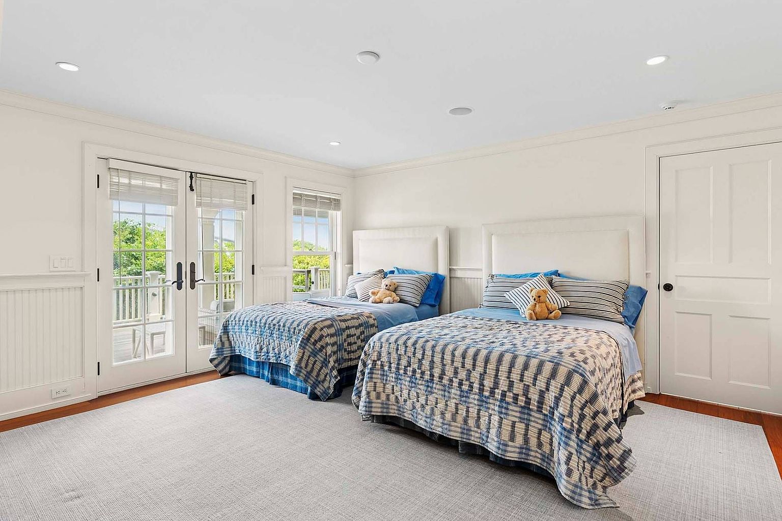 This is a bright and airy guest bedroom featuring two twin beds, each with a white upholstered headboard and blue and white patterned bedding. French doors lead to a balcony, enhancing the room's appeal. The walls are painted white, and the flooring is hardwood, covered by a light gray rug, creating a clean and inviting space.