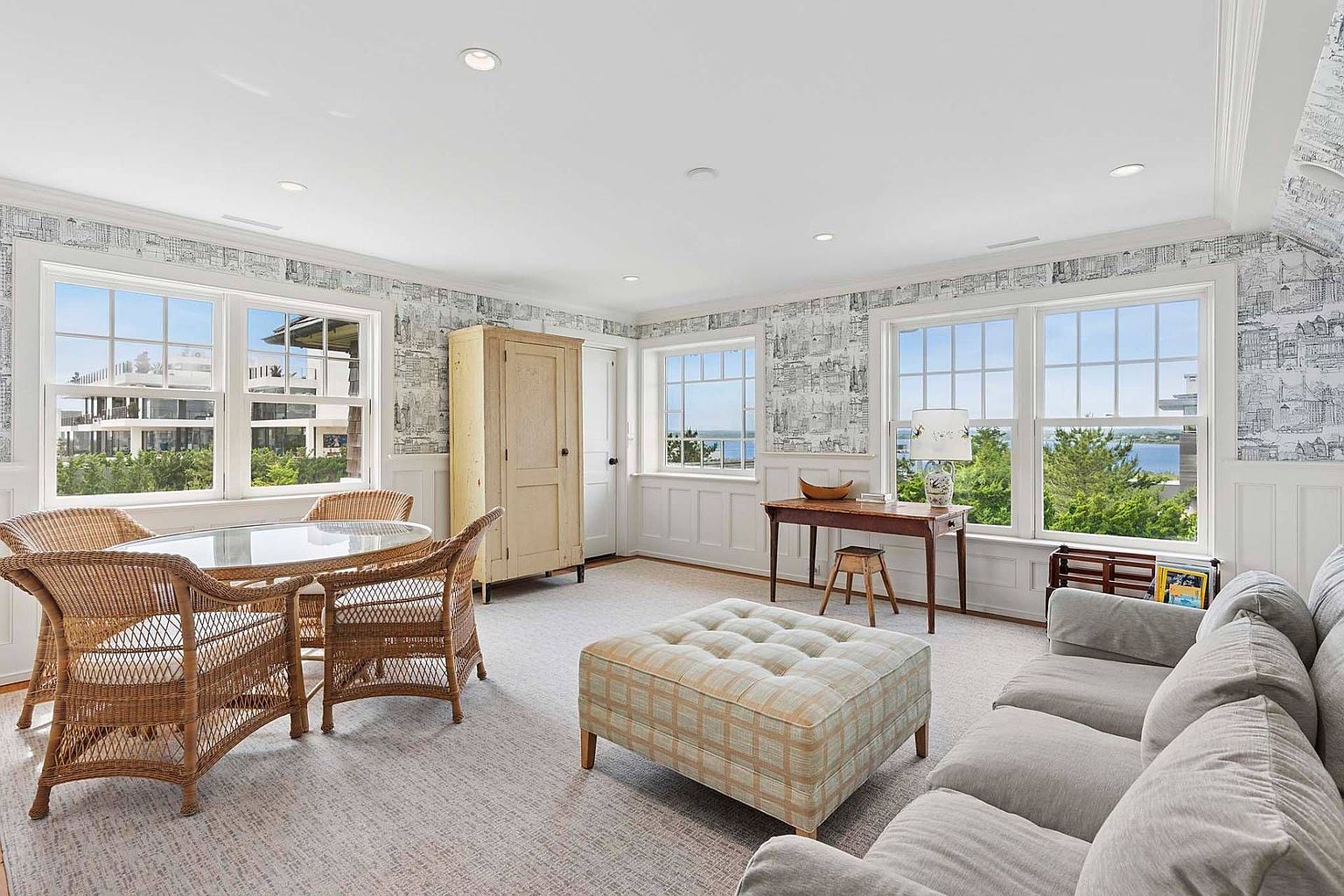 This is a bright and airy living room featuring a neutral color palette and ample natural light. The room includes a wicker dining set, a comfortable gray sofa, and a plaid ottoman, creating a cozy and inviting atmosphere. The walls are adorned with a unique wallpaper, and large windows offer pleasant views of the outdoors.