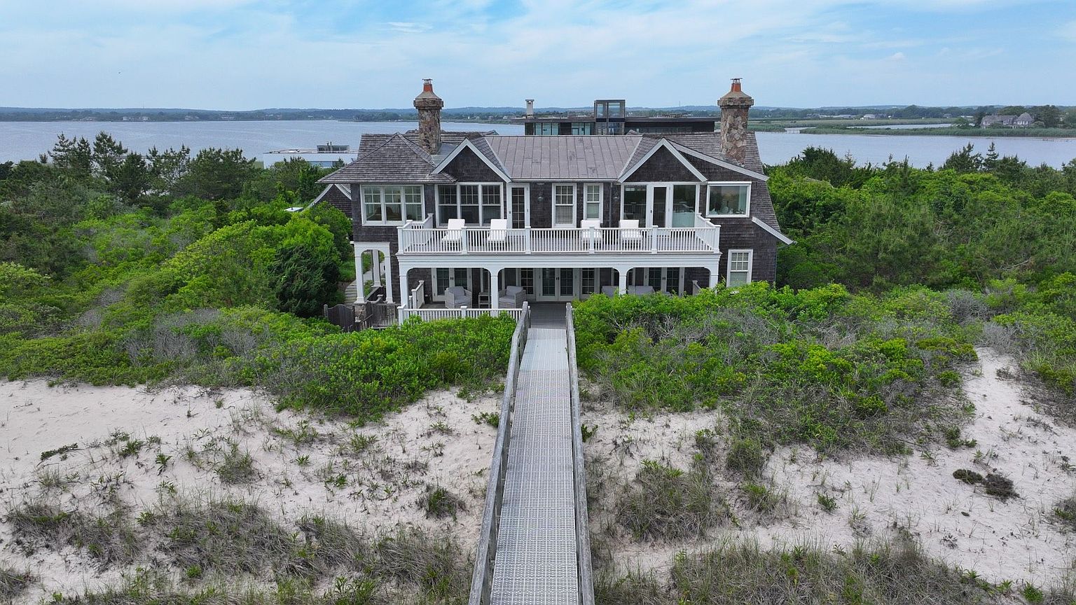 This aerial shot showcases a grand, two-story house nestled amidst lush greenery and sandy dunes, offering a sense of secluded luxury. The house features a large balcony, stone chimneys, and a walkway leading from the house to the beach, emphasizing its waterfront location and private access. The overall impression is one of an exclusive coastal retreat.