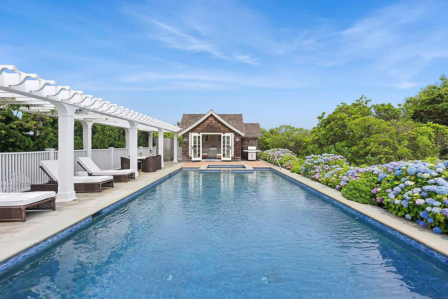 This image showcases a luxurious outdoor pool area. The rectangular pool is surrounded by a patio with lounge chairs and a pergola on one side, and a lush garden with hydrangeas on the other. A charming pool house stands at the far end, creating a serene and inviting backyard oasis.