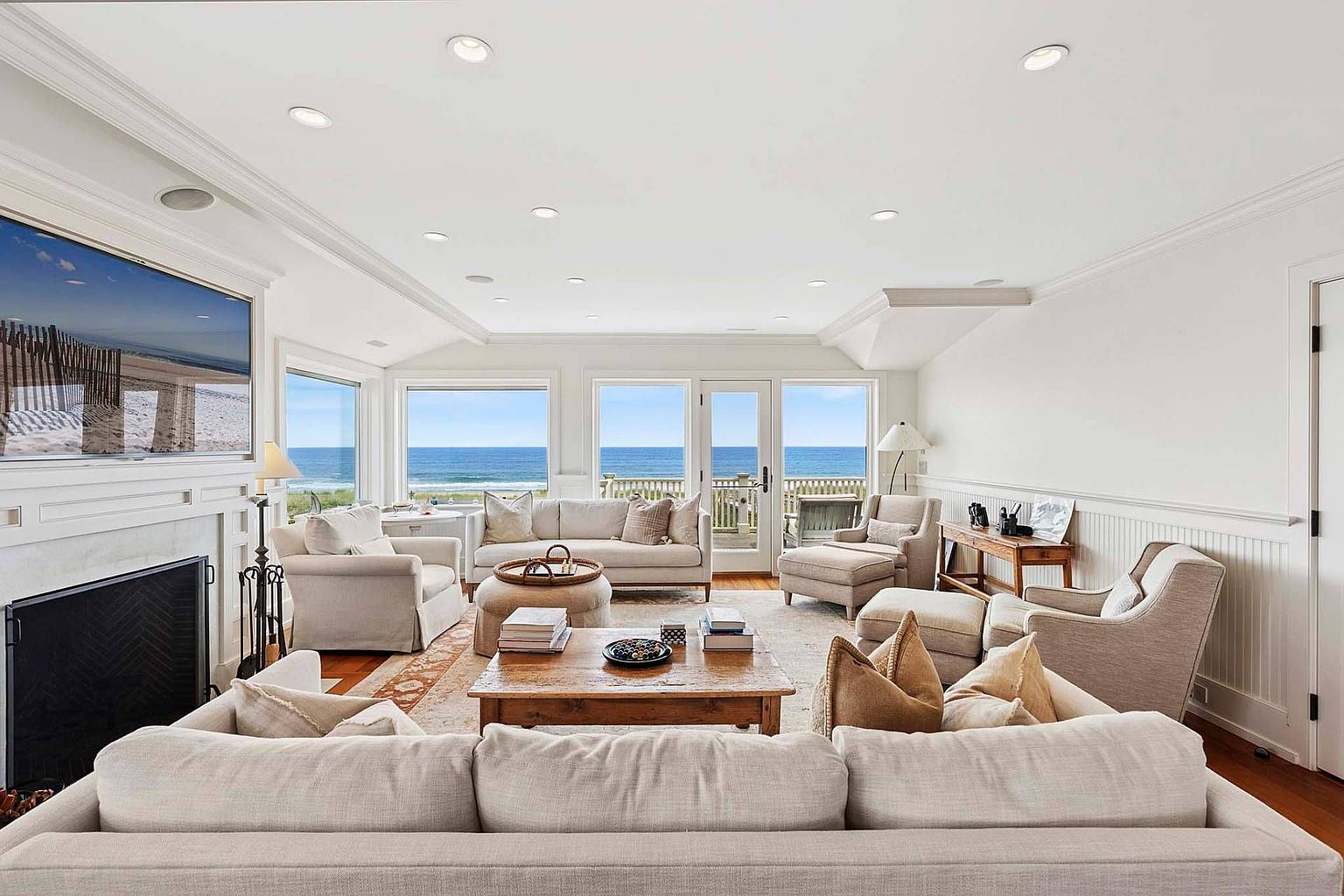 This is a bright and airy living room with a coastal aesthetic, featuring comfortable neutral-toned sofas and armchairs arranged around a wooden coffee table. Large windows offer stunning ocean views, enhancing the room's serene atmosphere. A fireplace with a mounted television adds a focal point, while the hardwood floors and white walls contribute to the room's clean and inviting feel.