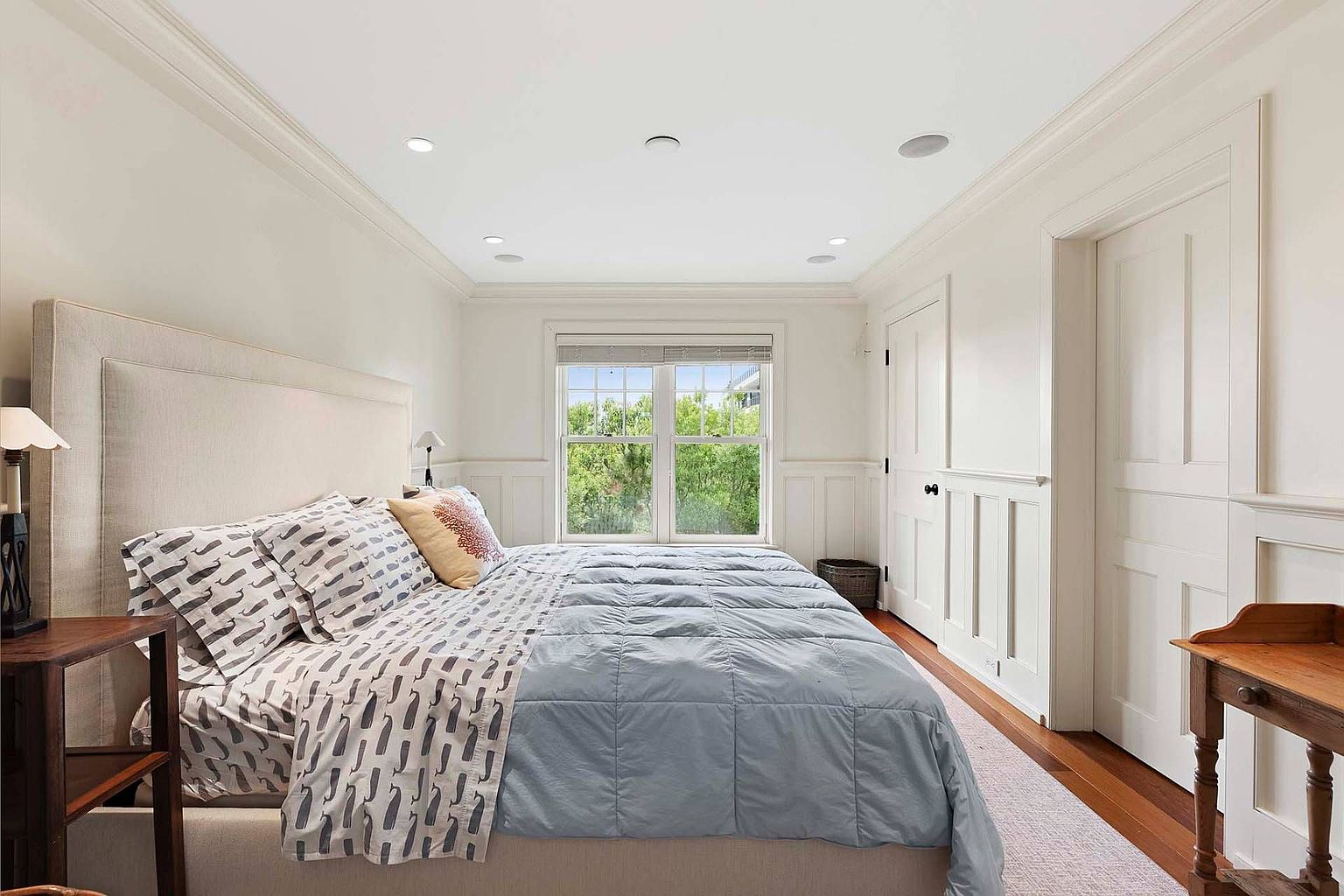 This is a well-lit primary bedroom featuring a large bed with a neutral-toned headboard and patterned bedding. The room has white walls with wainscoting, a window providing natural light, and hardwood floors partially covered by a light-colored rug. The overall impression is clean, comfortable, and inviting.