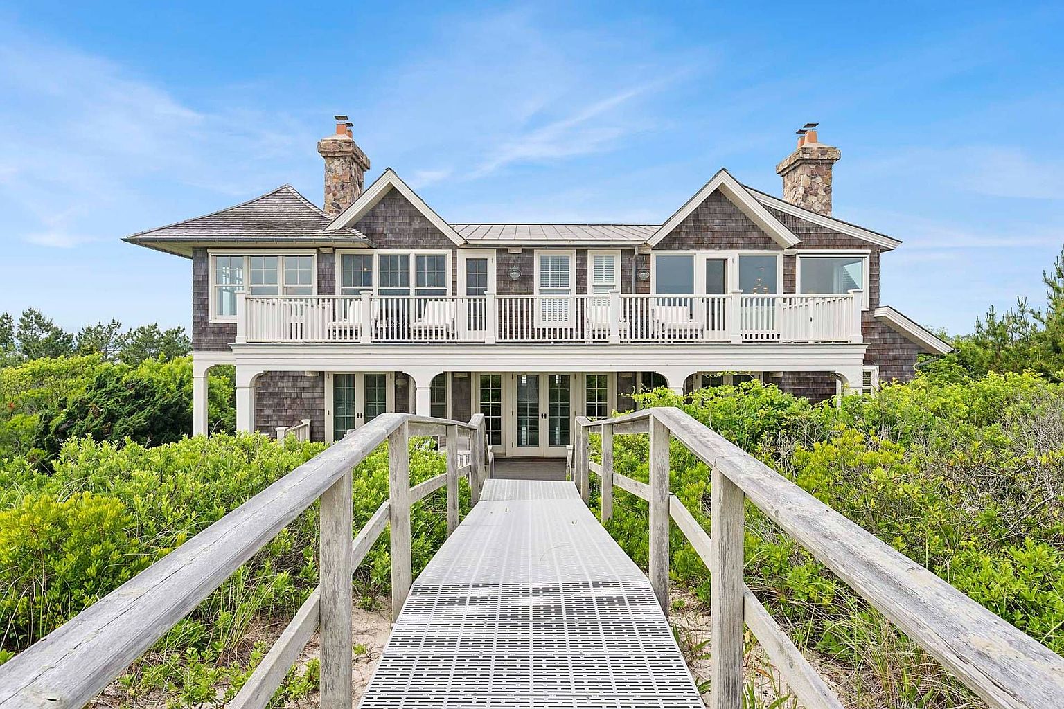 This is a front view of a large, luxurious beach house. The house features a shingle exterior, multiple windows, and a spacious balcony with white railings. A wooden walkway leads to the entrance, surrounded by lush greenery, creating a sense of privacy and tranquility.