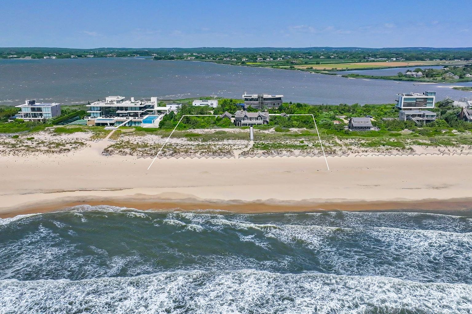 This aerial view showcases a stunning beachfront property with direct access to the ocean. The house is nestled among dunes and vegetation, with a defined property line extending to the sandy beach. The ocean waves create a dynamic foreground, while the landscape in the background adds depth and context to the location.