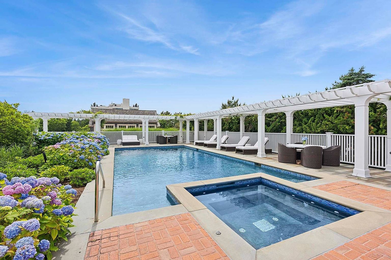 This image showcases a luxurious outdoor pool and spa area. The pool features a rectangular design with a separate hot tub, surrounded by a brick patio and a white pergola. Lounge chairs and a dining set are arranged along the pool deck, creating an inviting space for relaxation and entertainment.