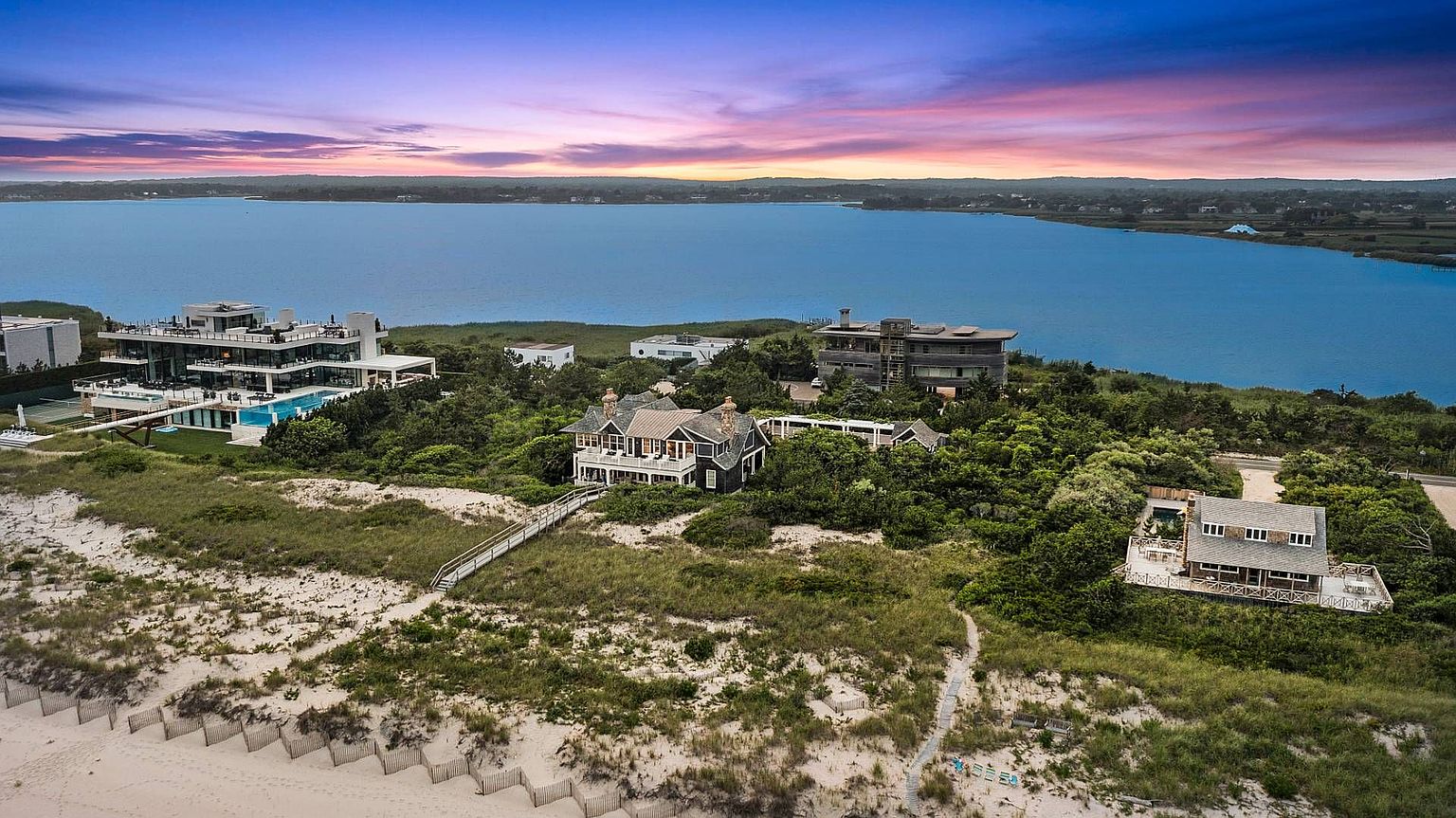 This aerial view showcases a collection of luxury homes nestled along a coastline, offering stunning waterfront views. The properties feature modern architectural designs, lush landscaping, and private beach access, creating an exclusive and serene living environment. The scene is set against a backdrop of a vibrant sunset sky and calm blue waters.