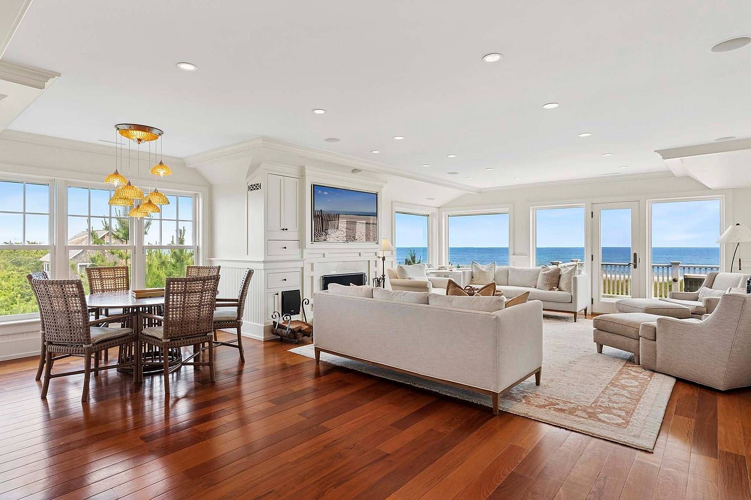 This is a bright and spacious living room with hardwood floors and large windows offering ocean views. The room features a dining area with a unique chandelier, a fireplace with a TV above it, and comfortable seating arrangements including sofas and armchairs. The overall design is elegant and coastal, creating a relaxing and inviting atmosphere.