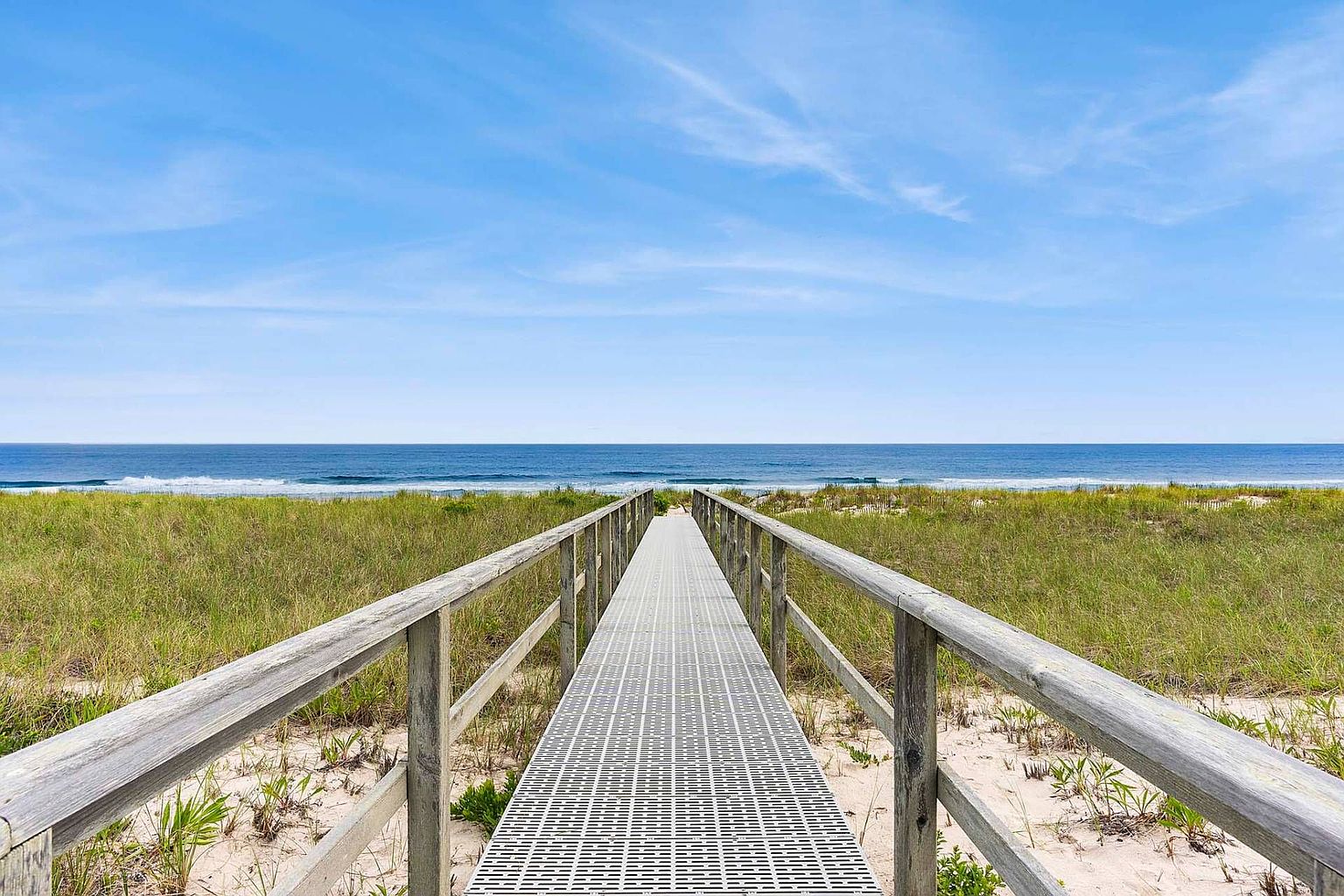 The image showcases a wooden boardwalk leading towards the ocean, flanked by grassy dunes on either side. The scene evokes a sense of tranquility and direct access to the beach, highlighting the property's proximity to the ocean. The clear blue sky and calm ocean waves enhance the appeal of this coastal setting.