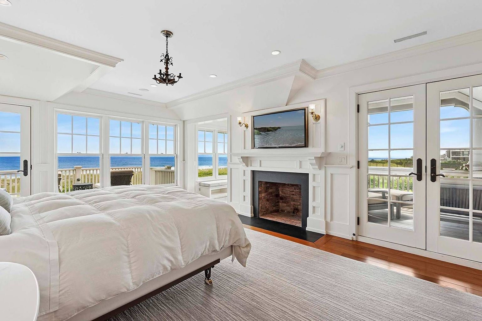 This is a bright and airy primary bedroom with a stunning ocean view. The room features a large bed with a white comforter, a fireplace with a mounted television, and access to a balcony through French doors. The decor is elegant and coastal, creating a relaxing and luxurious atmosphere.