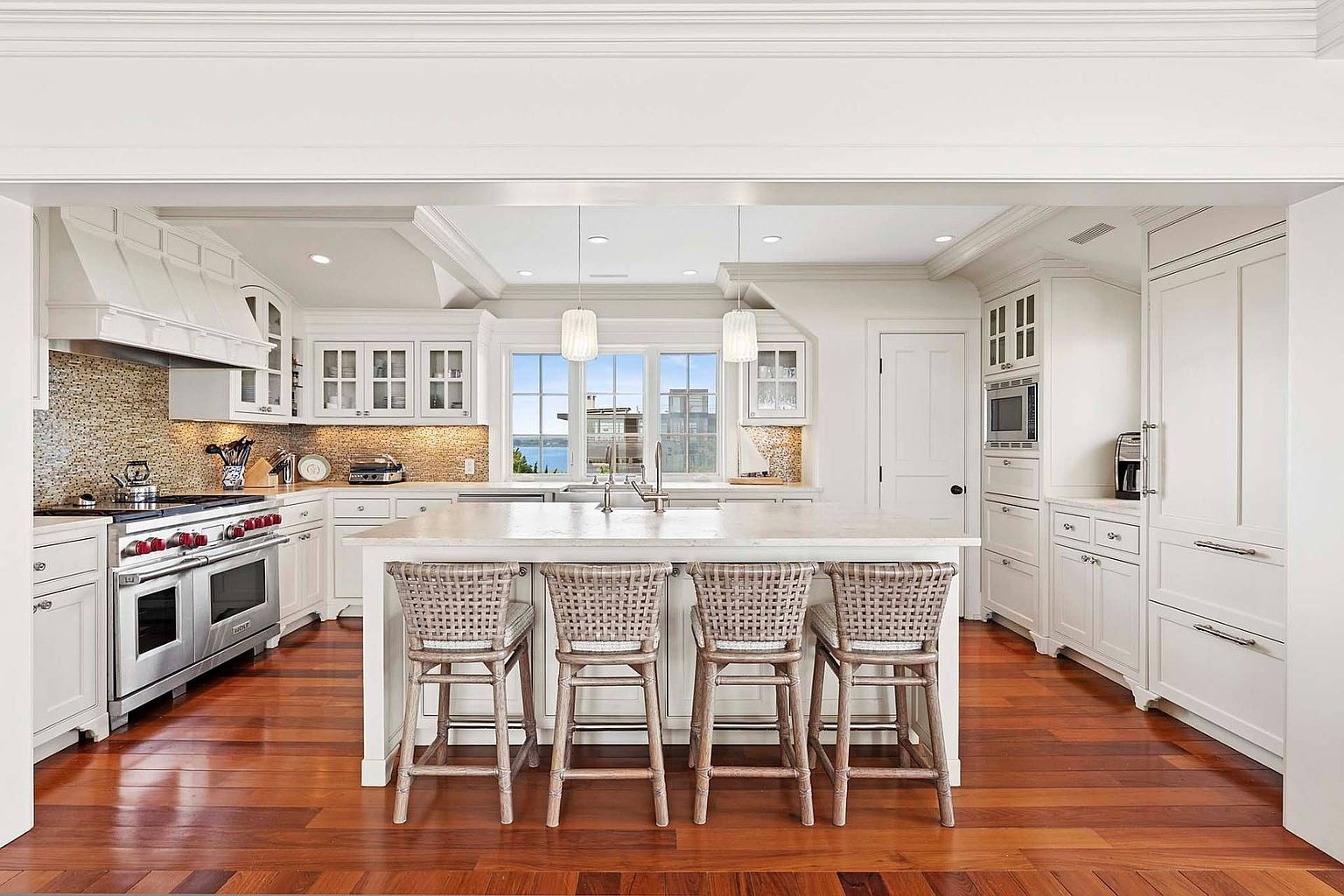 This is a bright and spacious kitchen featuring white cabinetry, a large center island with seating, and stainless steel appliances. The kitchen has hardwood floors and is well-lit with natural light from the windows above the sink and pendant lighting. The overall style is elegant and inviting, perfect for a modern home.