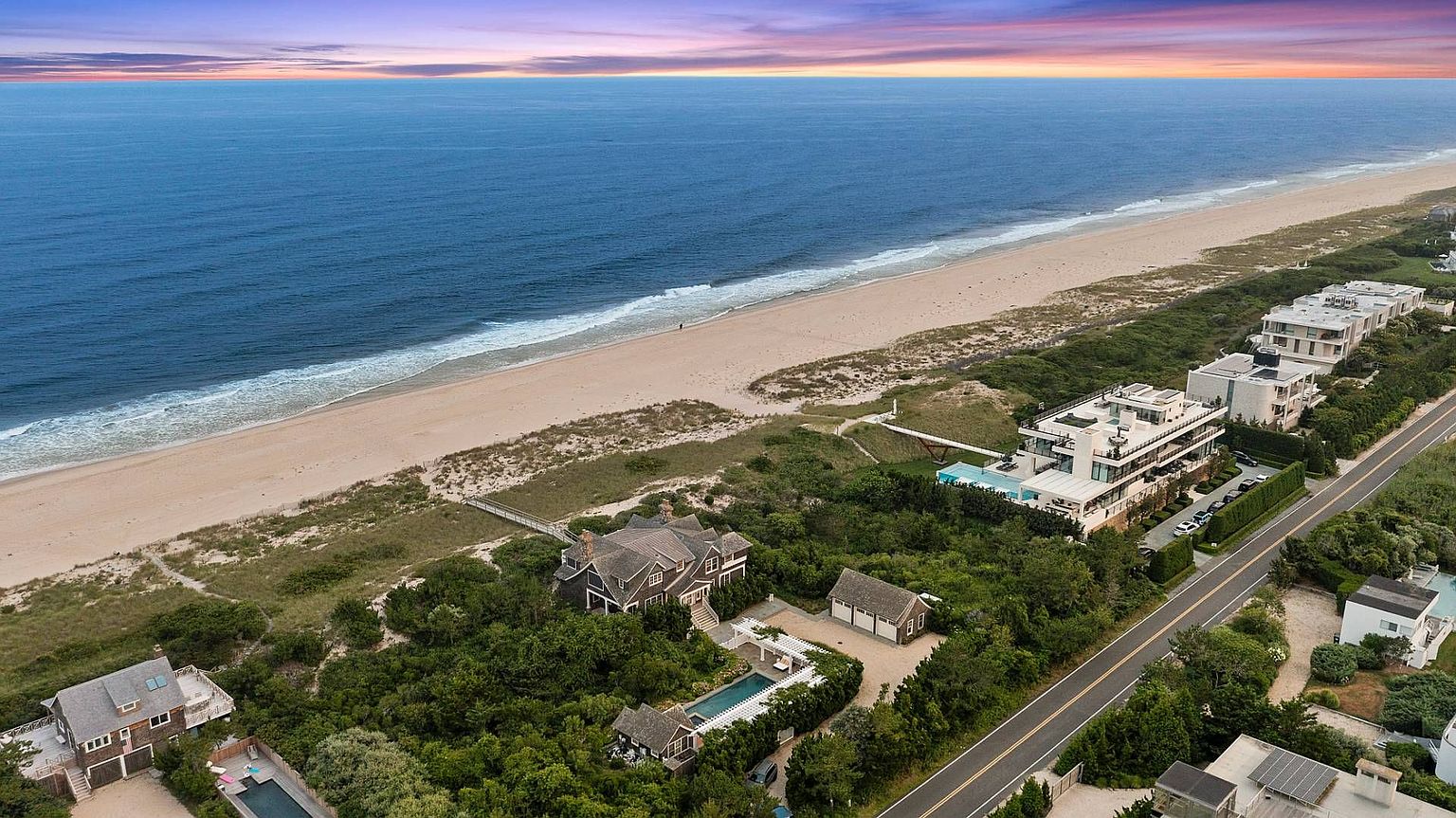 This aerial view showcases a stunning beachfront property with multiple buildings, including a main house, guest house, and garage. The property features a pool, lush landscaping, and direct access to a pristine sandy beach and the ocean. The perspective is a high-angle shot, emphasizing the property's location and luxurious amenities.