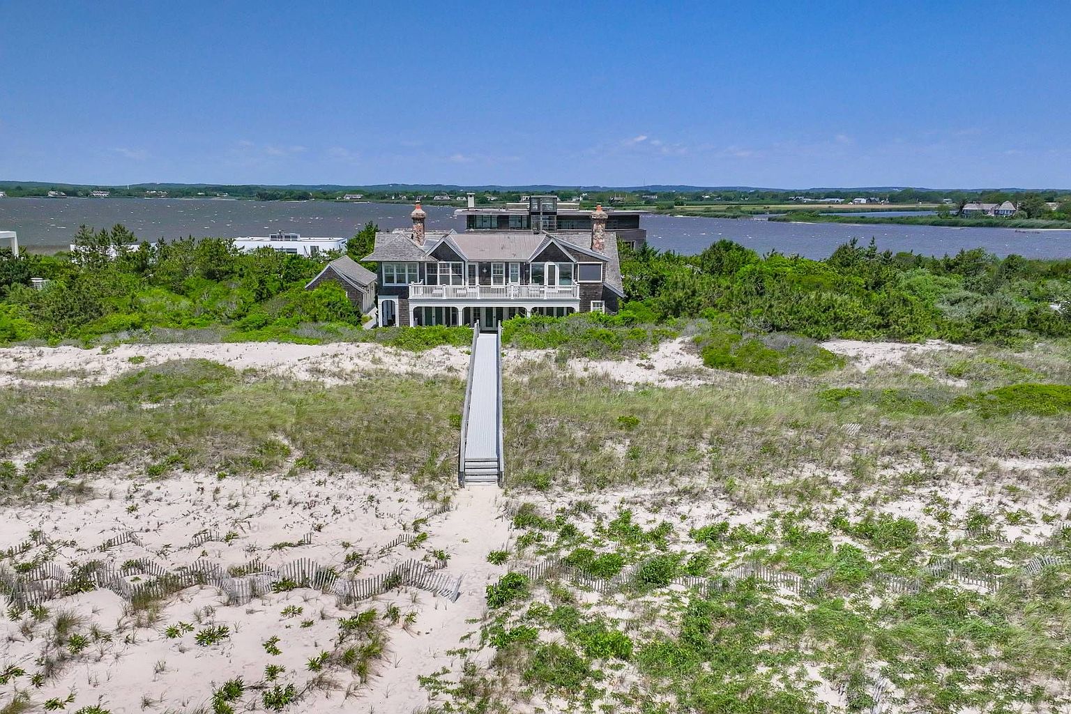 This aerial view showcases a stunning beachfront property with a large, multi-story house nestled among lush greenery. A wooden walkway leads from the house down to a sandy beach, with the ocean visible in the background. The house features a balcony and multiple chimneys, exuding a sense of luxury and privacy.