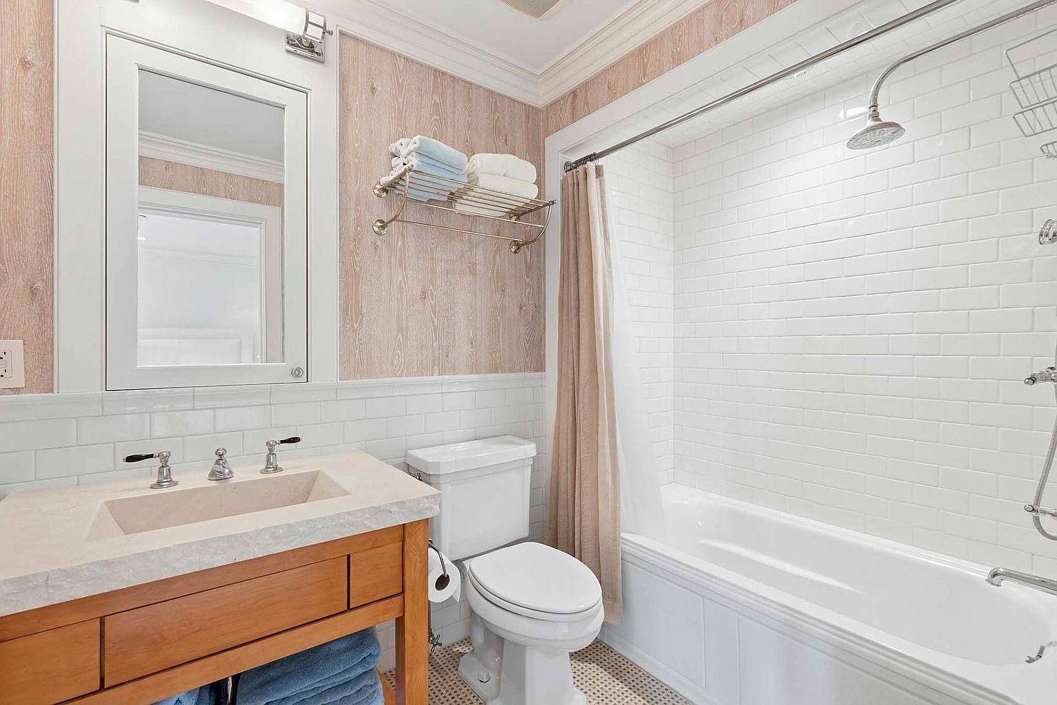 This is a well-lit bathroom featuring a wooden vanity with a stone countertop and a rectangular sink. A white-framed mirror hangs above the vanity, and a toilet is positioned next to it. The shower/tub area is enclosed with a shower curtain and features white subway tiles, creating a clean and classic look.
