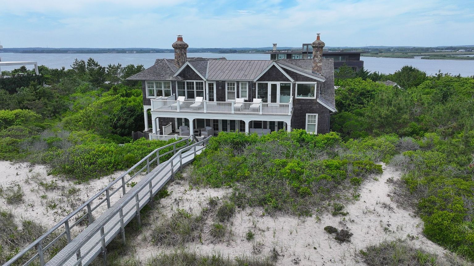 This aerial view showcases a stunning two-story home nestled amidst lush greenery and sandy dunes, likely near a beach. The house features a gray shingle exterior, a metal roof, and a spacious balcony with white railings, offering picturesque views of the water in the background. A wooden walkway leads to the house, adding to its secluded and luxurious appeal.