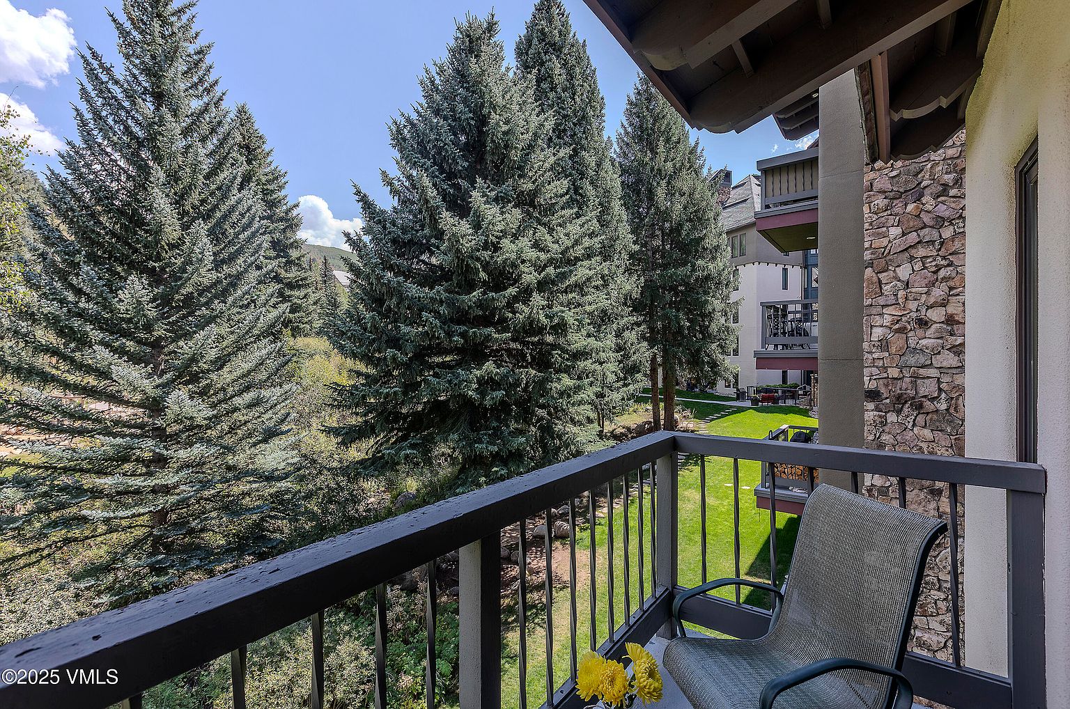 This image showcases a serene balcony view, featuring lush evergreen trees and a well-maintained lawn. The balcony itself has a dark railing and a comfortable chair, suggesting a relaxing outdoor space. The building's exterior combines stone and siding, adding architectural interest.