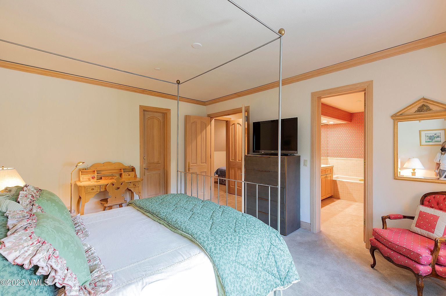 This is a primary bedroom featuring a four-poster bed with a light green patterned comforter and decorative pillows. The room includes a wooden desk, a TV on a dresser, and doorways leading to other rooms, including a bathroom with red wallpaper. The overall impression is a traditional and comfortable space.